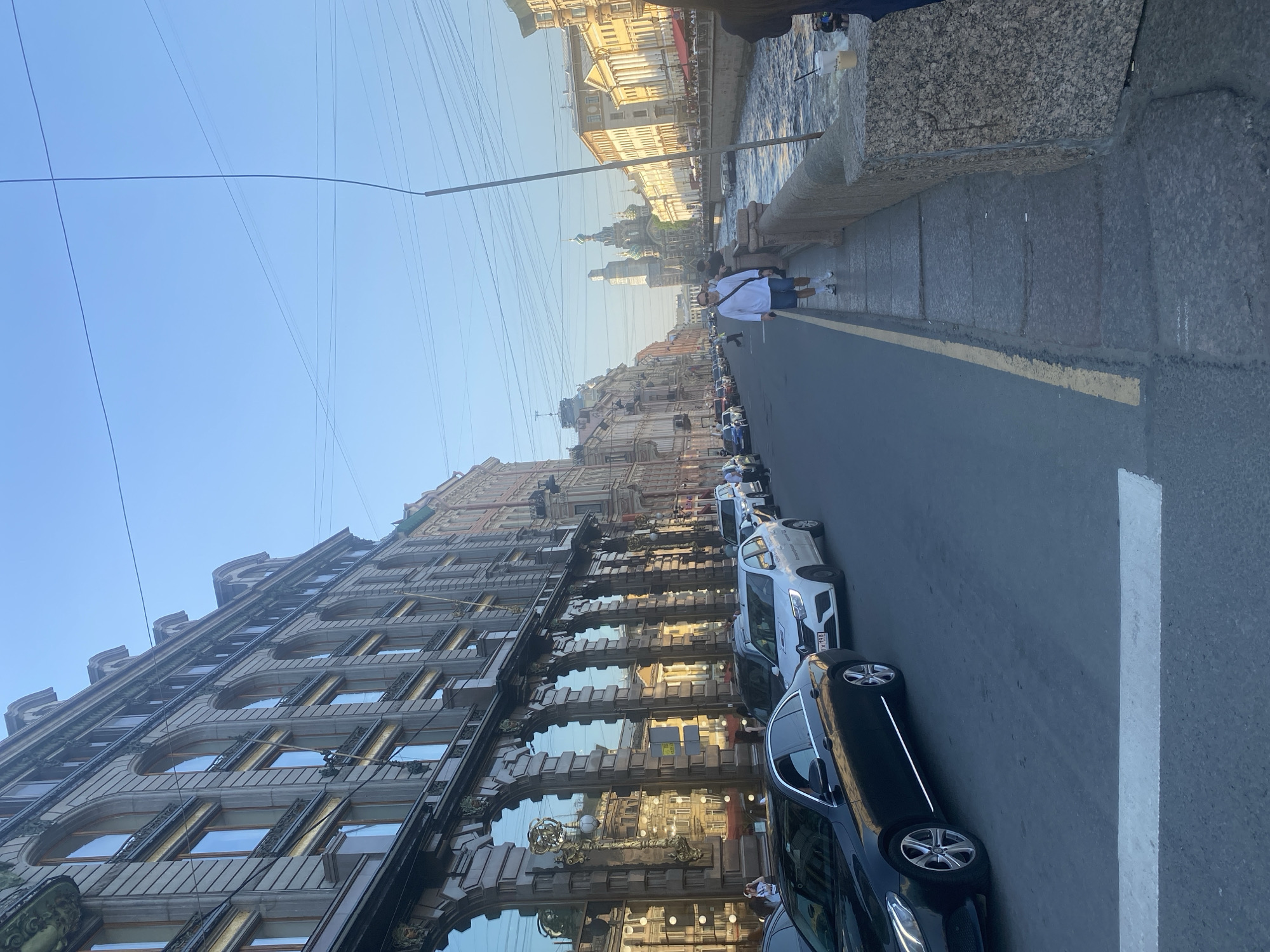 The image depicts a street scene in Saint Petersburg, Russia. A row of parked cars lines the right side of the street, adjacent to a pedestrian walkway bounded by a stone embankment. Across the street, a large building with ornate facade details, including arched windows and decorative metalwork, rises. Reflections in the building's windows show the buildings across the canal. A few pedestrians are walking along the embankment. In the distance, the distinctive spires of the Church of the Savior on Spilled Blood are visible. Overhead, numerous electrical wires stretch across the sky. The environment is bright, indicating daylight, with a clear sky.