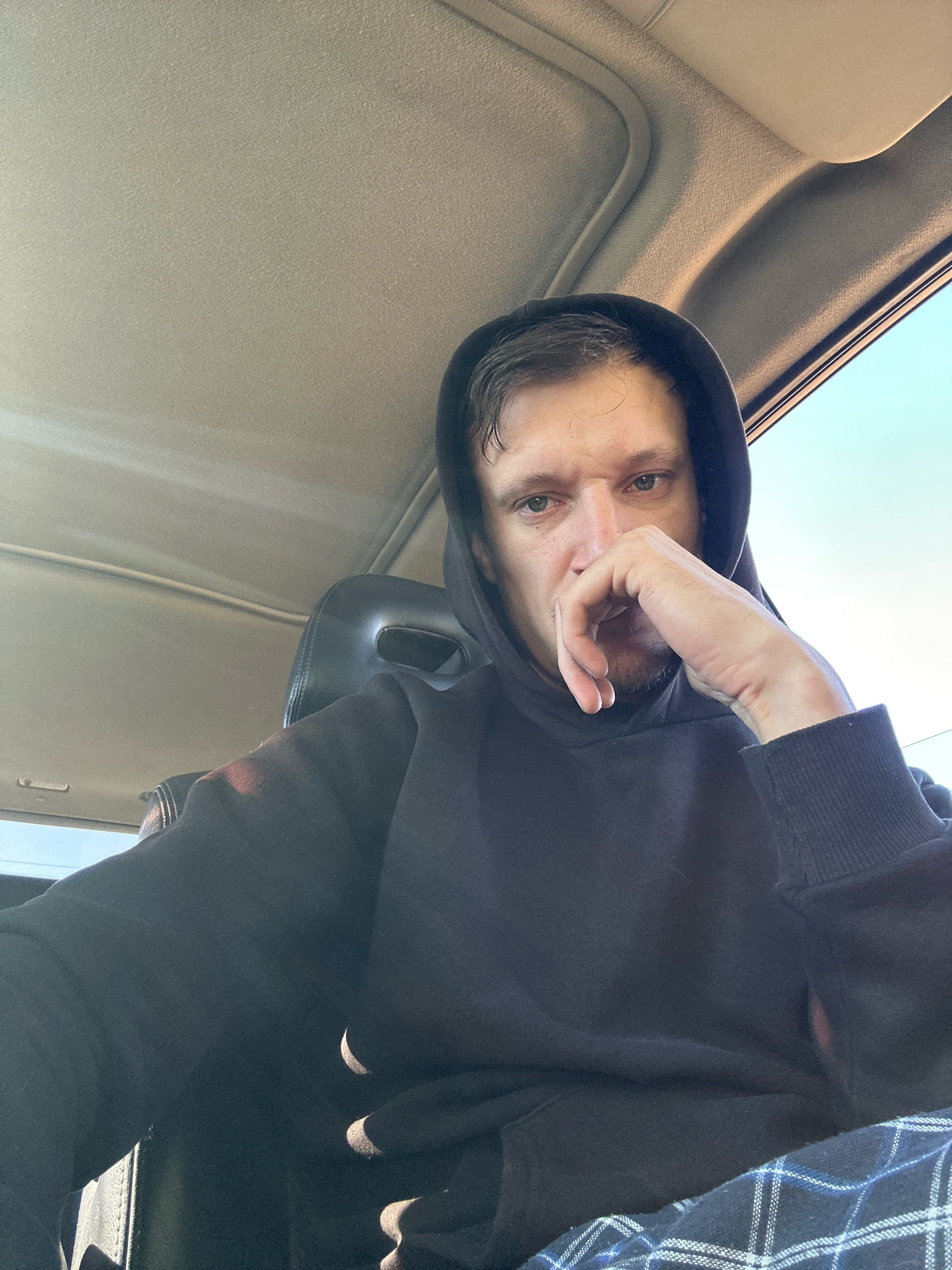 A man, wearing a black hooded sweatshirt and plaid pants, is captured in a close-up, front-facing selfie. His left hand rests on his chin, and his gaze is directed forward. The scene appears to be inside a car, with the textured beige interior of the car's roof visible above him. Sunlight streams in from the right side of the frame, illuminating the interior and casting shadows. The background is partially obscured by the car's seat. The image does not provide any specific details about the city of Makiv, Ukraine, or any contextual information that would place it in that location.
