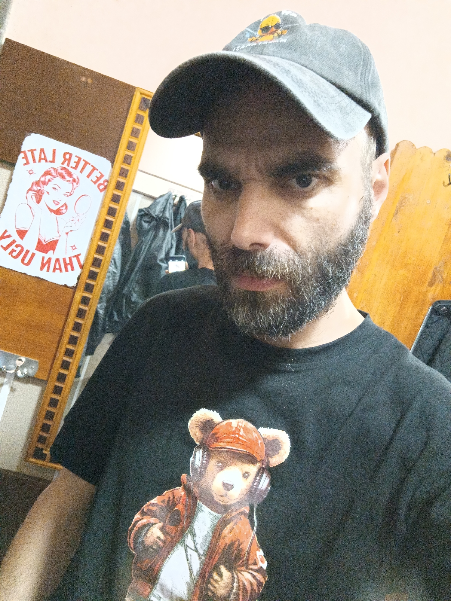 A man with a beard and a grey baseball cap is taking a selfie in what appears to be a room with a mirror. He is wearing a black t-shirt with a teddy bear graphic. In the mirror's reflection, we can see part of the man taking the photo, another reflection, a black jacket, and a poster that reads "BETTER LATE THAN UGLY." The background consists of wooden paneling and a light-colored wall.
