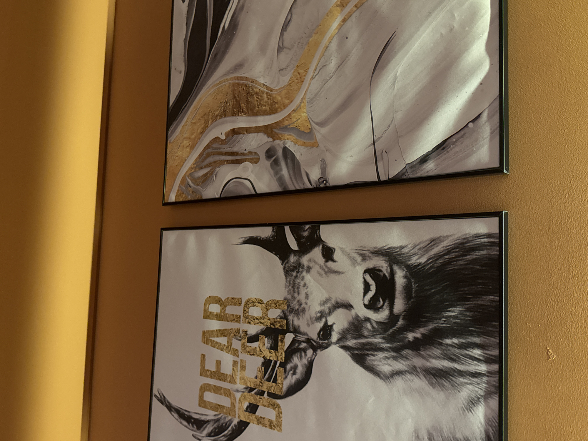 Two framed art pieces hang on a mustard-yellow wall. The top piece is an abstract design in white, gray, black, and gold. The lower piece is a black-and-white image of a deer with the word "DEAR" written vertically in gold, overlaying part of the deer's head. The location is likely indoors. Đà Nẵng, Vietnam, is not identifiable from the image.