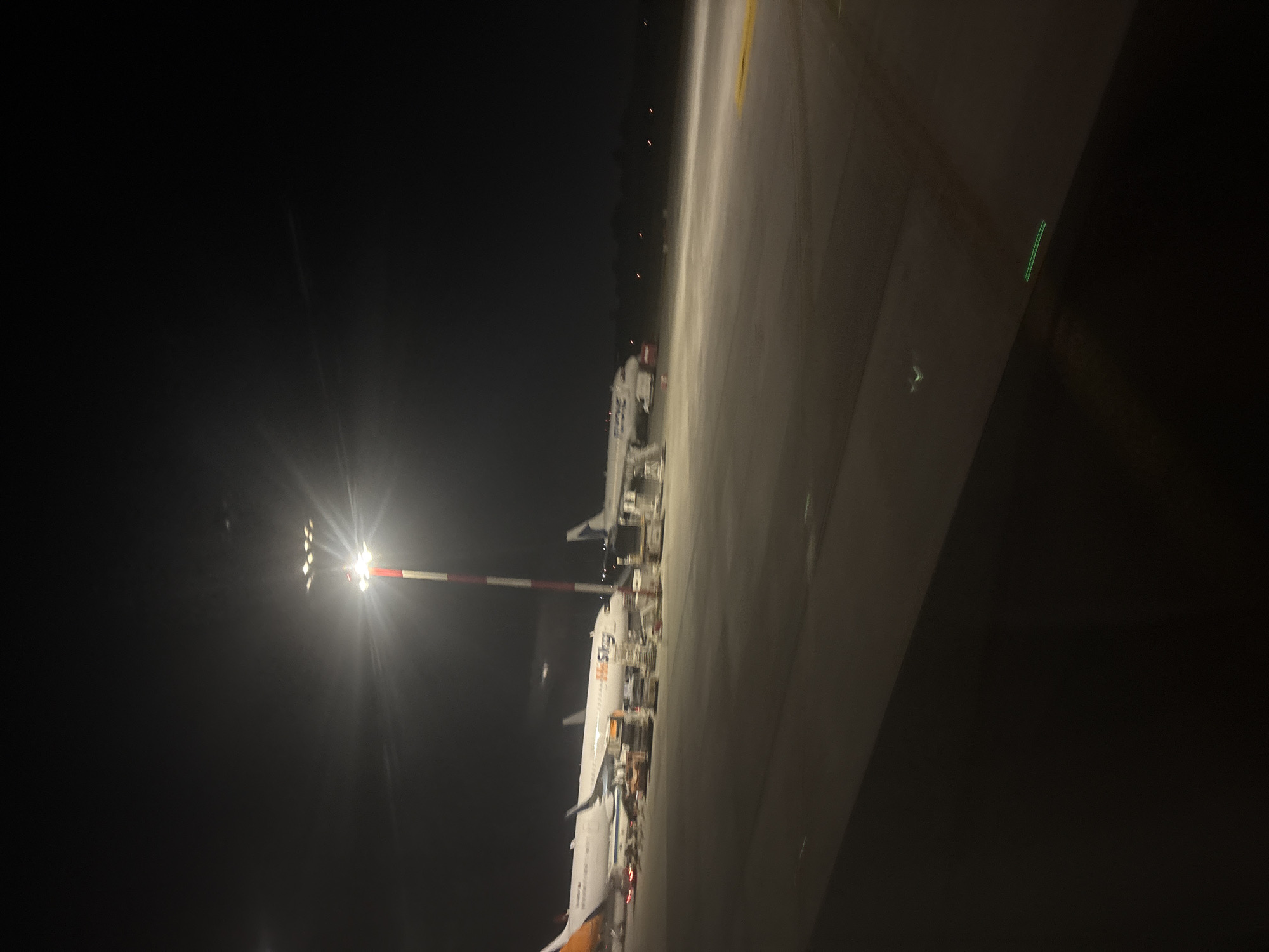 The image depicts an airport tarmac at night in Otopeni, Romania, likely at Henri Coandă International Airport. A bright floodlight with a red and white pole illuminates the scene, casting long shadows. Two airplanes are parked on the tarmac. The closer airplane, on the left, is a white passenger jet with orange and yellow markings on its tail and appears to be branded "Pegasus." It is equipped with stairs and what appear to be baggage carts and other ground support equipment. Further to the right, another white passenger jet is visible, with the visible portion of its livery being blue and white. The tarmac itself is paved and appears somewhat damp or dusty. In the background, the dark sky is sparsely dotted with distant lights, suggesting the airport is operational. The foreground shows a dark, slightly reflective surface, possibly a window or a vehicle's surface, from which the photo was taken.