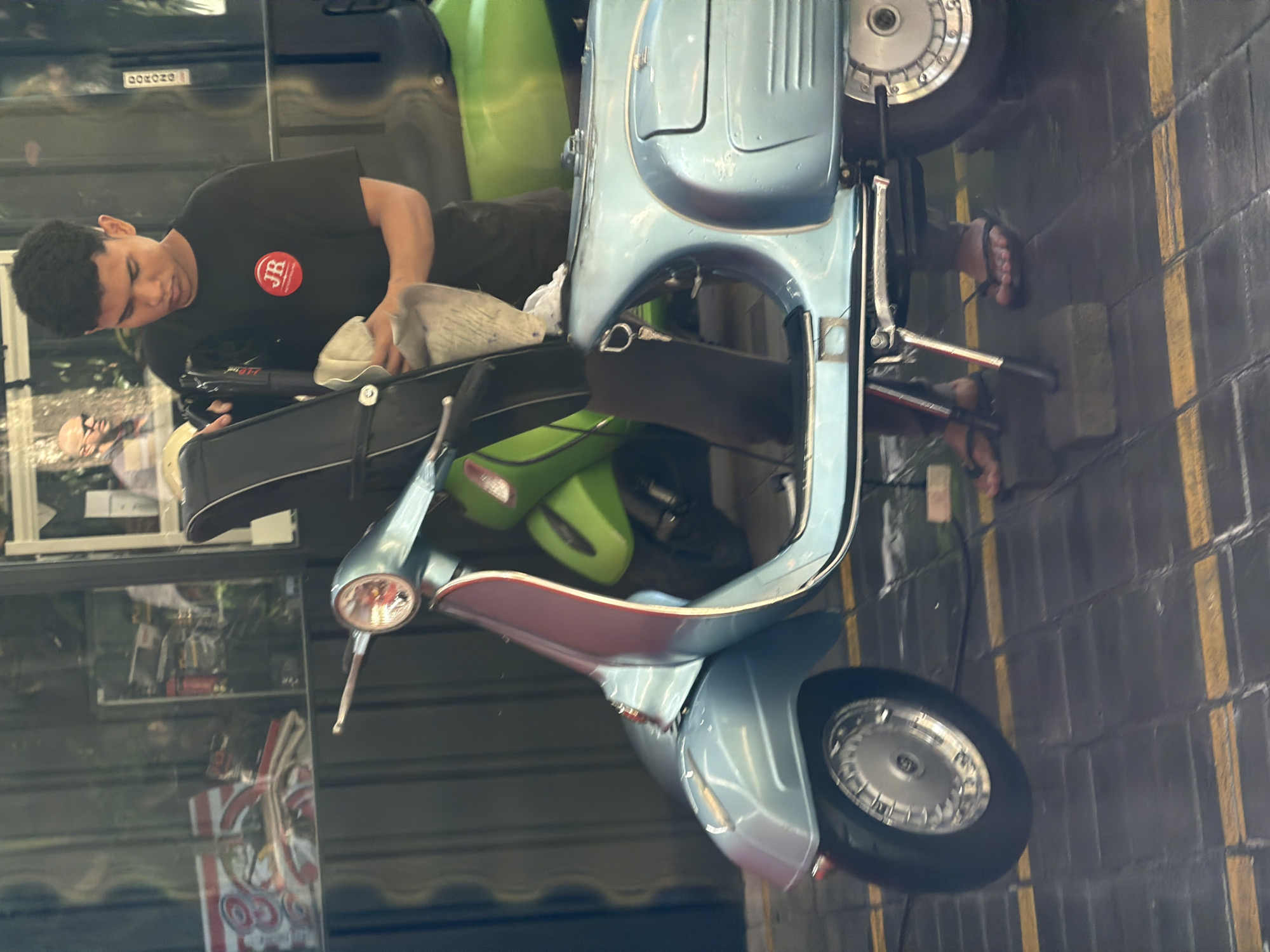 In Kuta, Indonesia, a young man wearing a black t-shirt with a red "JR" logo is polishing a light blue Vespa scooter. The scooter is parked on a wet, tiled surface marked with yellow lines. Another green scooter is partially visible behind the blue one. The background shows a shop with windows displaying various items, including what appears to be vintage motorcycle parts and merchandise. Reflections in the window suggest an indoor environment, possibly a repair shop or a retail store. The man is using a light-colored cloth to clean the scooter.