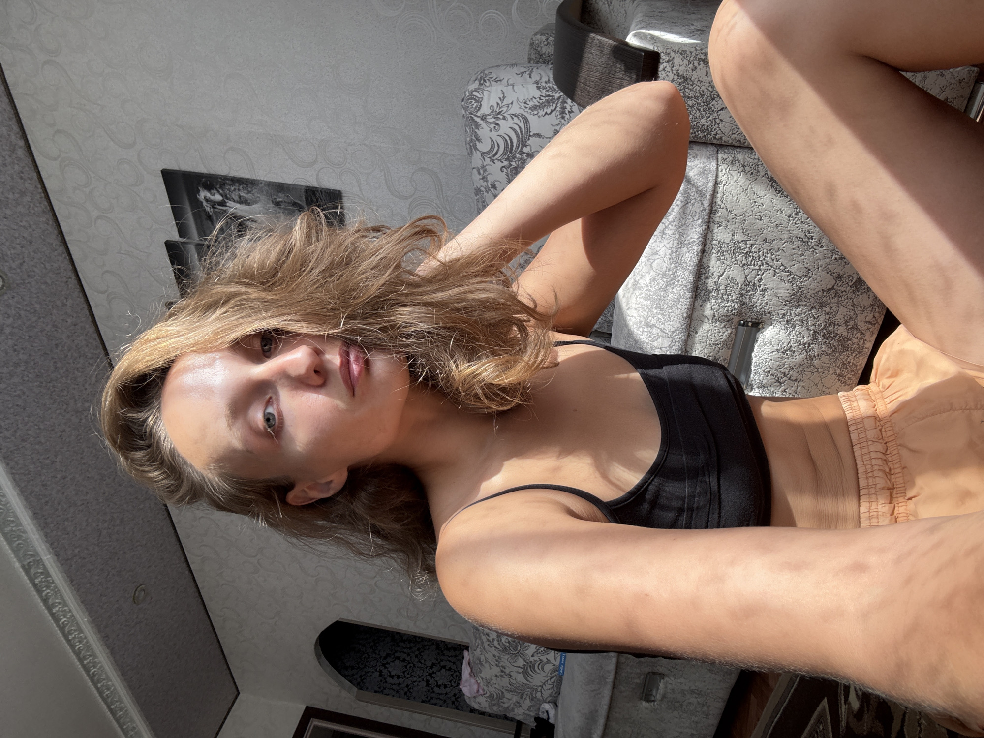 A young woman with light brown hair is sitting on the floor in a room, possibly an apartment in Kuyeda, Russia. She is wearing a black sports bra and peach-colored shorts. Sunlight streams in from the side, casting shadows across her body and the patterned upholstery of a sofa behind her. Her left arm is bent, with her hand touching her hair, and her right arm is resting on her leg. The room appears to be furnished with a sofa and has patterned wallpaper. Two framed pictures hang on the wall behind her.