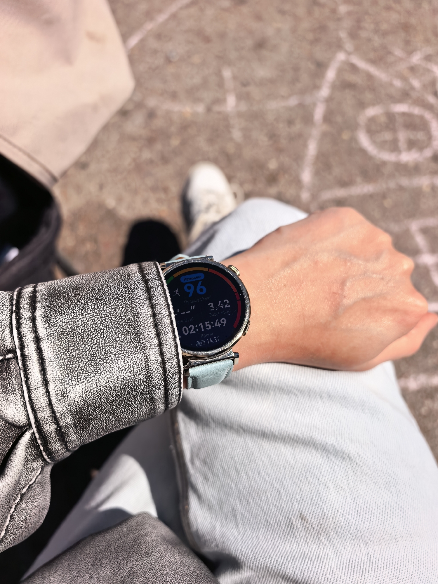 A person is seated outside on a concrete surface with chalk drawings visible. They are wearing light blue jeans and a silver-grey jacket. A smartwatch on their left wrist displays a screen showing a heart rate of 96, a distance of 3.42, and a time of 02:15:49. A white sneaker is visible in the background. The exact location is Sterlitamak, Russia, but there is no visual confirmation of that in the photo.