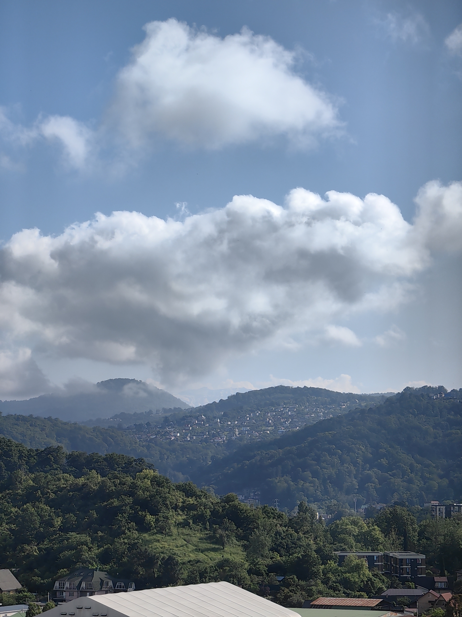 This is an aerial, wide-angle shot looking east-northeast over the city of Sochi, Russia. The image features a layered landscape beginning with a foreground of trees and buildings, transitioning to rolling, densely forested hills in the mid-ground, and culminating in distant, hazy mountains. The sky is a bright blue with large, white cumulus clouds that cast shadows on the landscape. The upper slopes of the mid-ground hills are dotted with a dense cluster of white-roofed buildings, indicating a residential area built on the incline. The lower slopes are covered in thick green forest. In the foreground, there are several modern apartment buildings and what appear to be commercial buildings with white roofs. The overall impression is of a sun-drenched, verdant city nestled within a mountainous region.