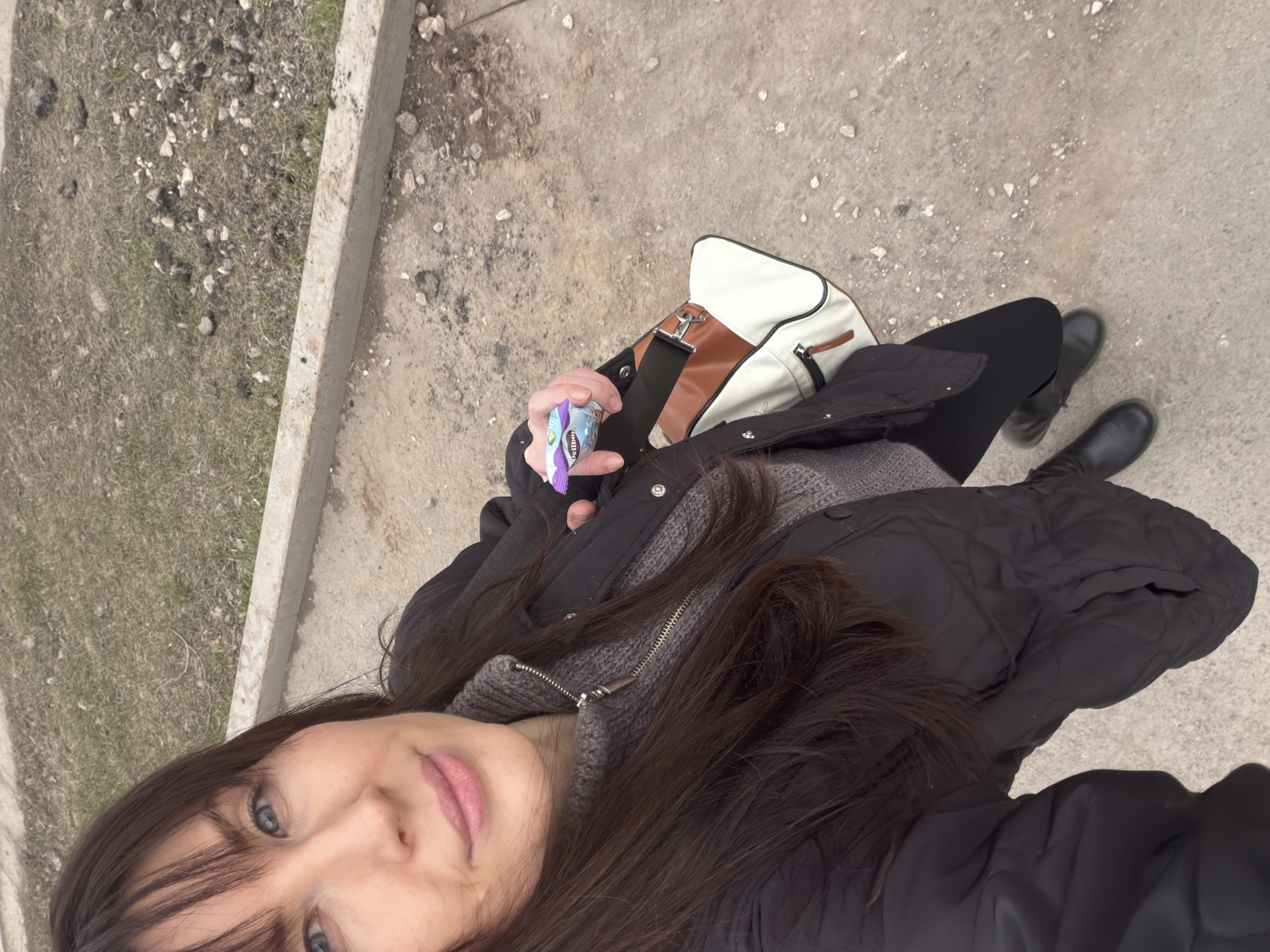 A selfie taken in Perm, Russia shows a person with long brown hair, wearing a gray sweater, black down jacket, and black leggings with black boots. A white, brown, and black purse is also visible. The person is holding a blue and purple wrapper. The location appears to be a sidewalk or concrete area near a grassy verge with sparse vegetation and rocks. The time of year appears to be late fall or early spring.