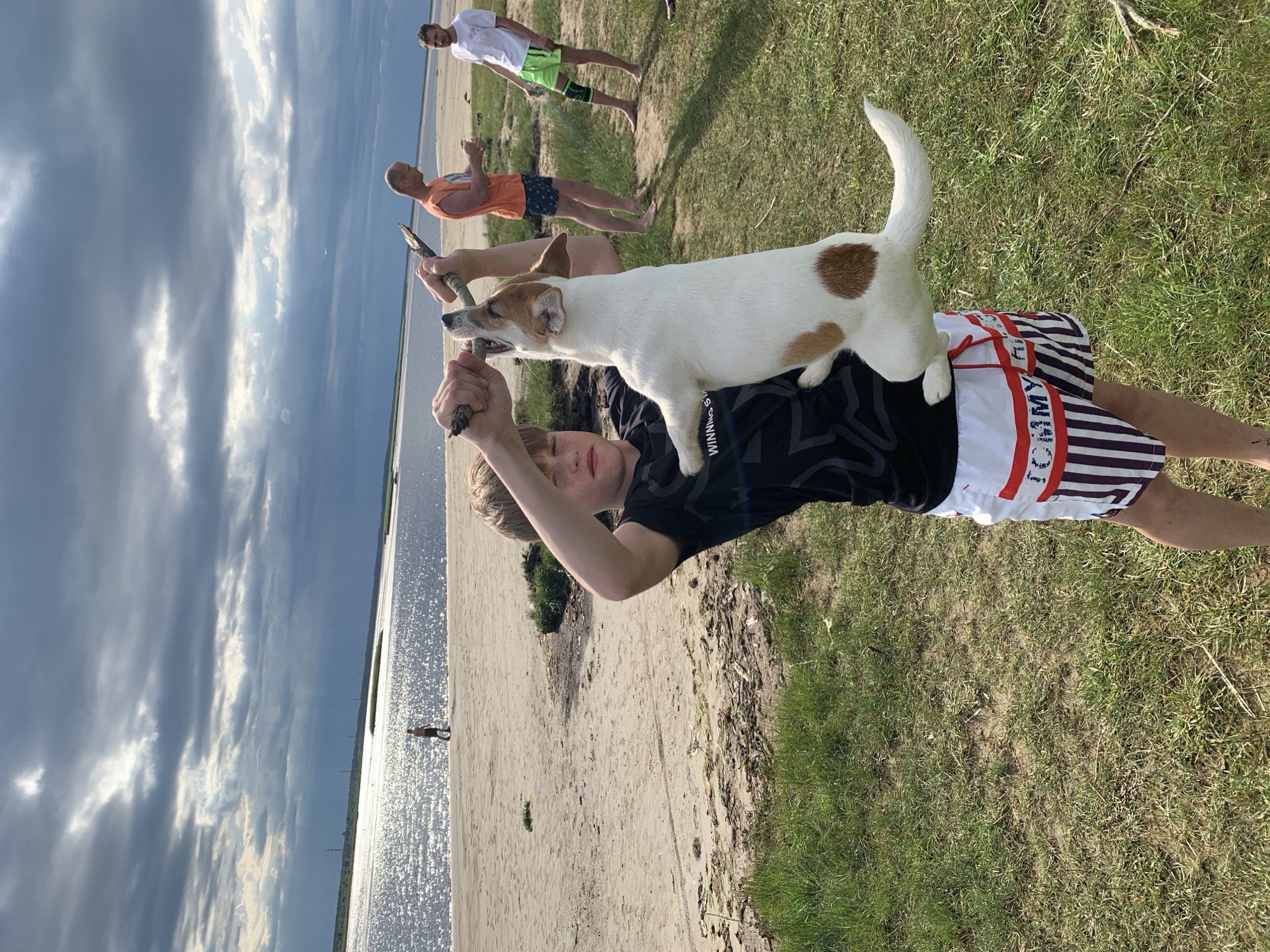 An outdoor, eye-level shot shows a young boy in the foreground holding a Jack Russell Terrier in the air by a stick. The boy is wearing a black t-shirt and white and red striped shorts with a black and purple striped pattern at the bottom. He has brown hair and is looking at the camera. The dog is white with brown patches and is gripping the stick in its mouth. The background is a sandy beach with a large body of water and a cloudy sky. There are two men in the background, one wearing an orange t-shirt and the other a white t-shirt and green shorts. The scene appears to be taking place on a beach in Severodvinsk, Russia.
