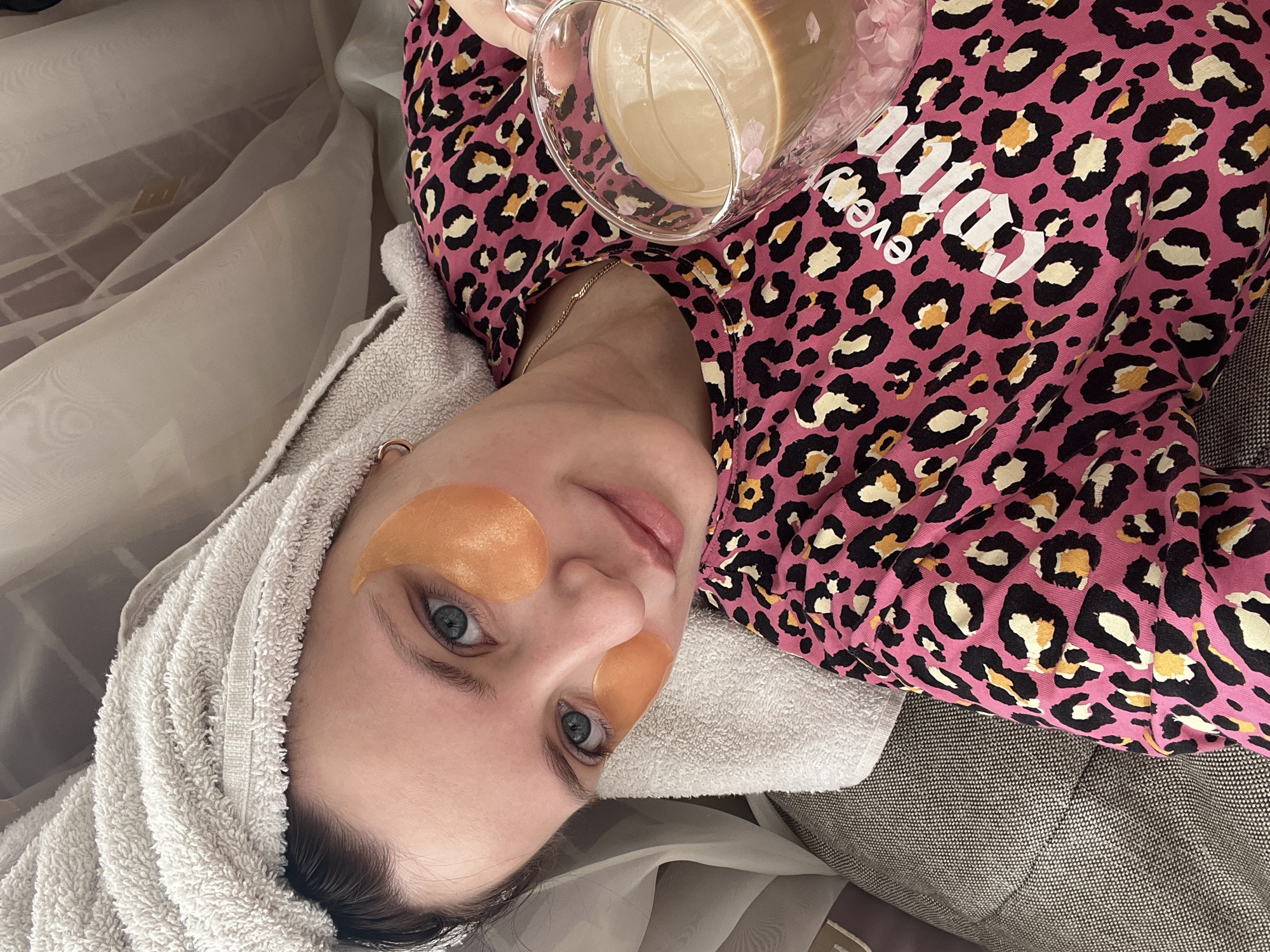 An indoor, eye-level shot shows a young woman relaxing, likely on a bed or couch. She lies on her back, gazing directly at the camera with a soft, content expression. Her light blue-green eyes are prominent, accentuated by golden-orange under-eye patches. A white terrycloth towel is wrapped around her head, suggesting a recent shower or a self-care routine.
She is wearing a vibrant pink long-sleeved top with a distinctive black, white, and yellow leopard print. Partially visible white text on her top reads "Everyd". Around her neck, a delicate gold chain necklace is visible. Her right hand holds a clear glass mug filled with a light brown liquid, resembling coffee or a latte.
The background features textured fabrics: a dark gray, subtly patterned material (possibly upholstery) is visible to the right and beneath her, while soft, translucent white drapes or curtains with a faint grid pattern are on the left. The scene evokes a sense of peaceful relaxation, possibly during a morning or evening routine in a domestic setting.