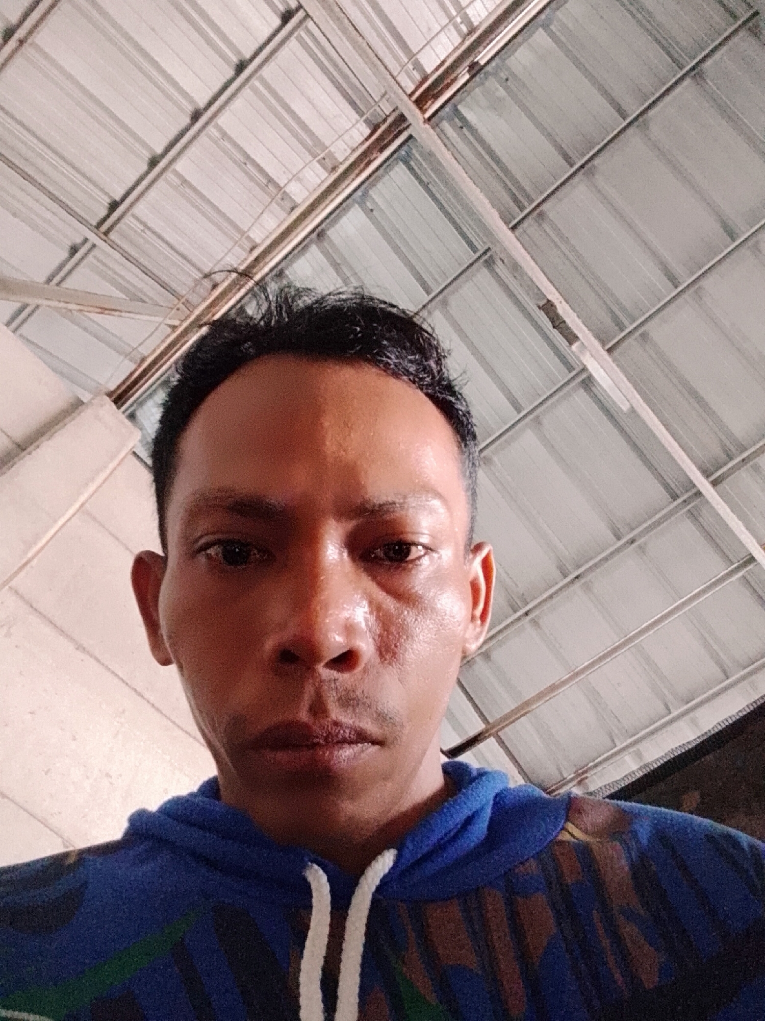 The image depicts a close-up view of a man's face taken from a low angle. He has short black hair and appears to be wearing a blue hoodie with white drawstrings. The man's skin appears slightly shiny, potentially indicating moisture or sweat. The background shows a ceiling constructed of corrugated metal sheets supported by a framework. The location is identified as Bukit Ketok, Indonesia. No specific actions or interactions are evident from the image.