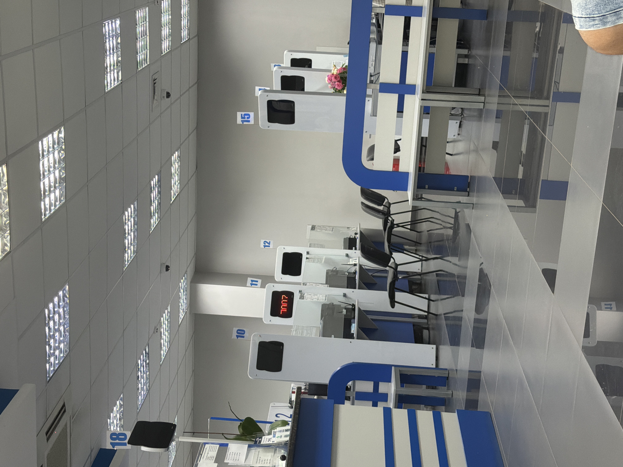 The image depicts the interior of a modern, well-lit administrative office or service center in Strășeni, Moldova. The room features a tiled floor, a suspended ceiling with fluorescent lighting panels, and light gray walls. Several service counters are visible, some equipped with digital display screens and computer terminals. Numbered signs (18, 10, 11, 12, 15) are mounted on the walls, indicating service stations or waiting areas. One digital display shows the number "7007". Chairs are arranged in front of some counters. A small bouquet of pink roses is placed on a counter. The overall environment suggests an organized and functional space designed for customer service or bureaucratic procedures.