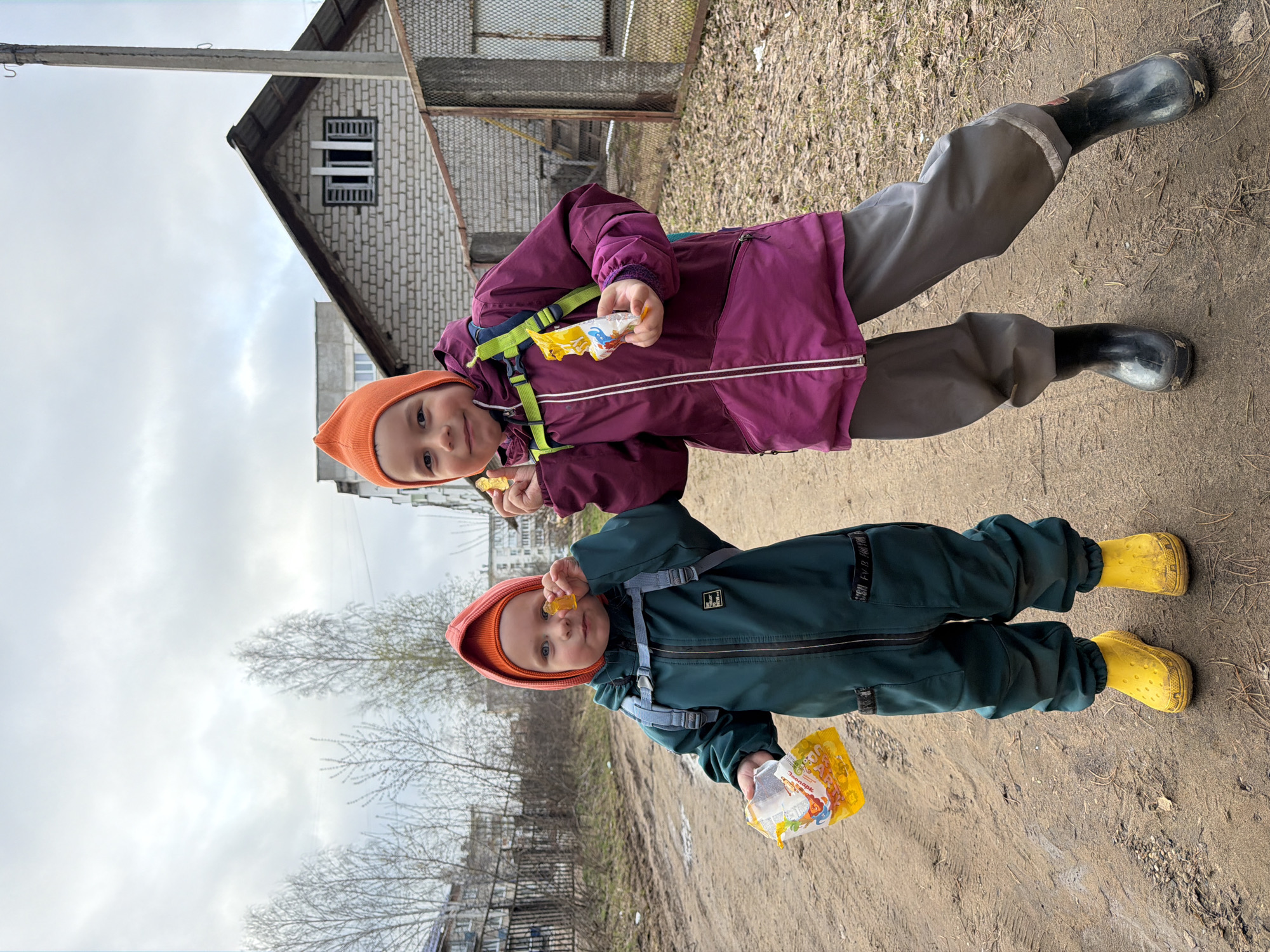 Two young children are captured in an outdoor setting, likely in Chaikovsky, Russia, on what appears to be a dirt path. The image is taken on an overcast day, suggesting cool weather, possibly in late winter or early spring, given the bare trees in the background.
The child on the right (taller) wears a purple hooded jacket, grey waterproof pants, black rubber boots, and a bright orange knit hat. They are holding a snack, possibly a juice pouch or similar food item, and appear to be eating something. The child on the left (smaller) is dressed in a dark green waterproof full-body suit (overalls), bright yellow rubber boots, and a matching orange knit hat. This child holds a yellow snack package and a small, translucent yellow piece of food. Both children are looking directly at the camera with pleasant, content expressions.
The ground beneath them is unpaved, appearing muddy and worn. In the background, a light-colored brick or block house with a dark sloped roof is visible, along with a utility pole and a metal fence. Further in the distance to the left, more structures and bare trees are present under the grey sky. The snack package held by the smaller child has visible Russian text, reading "Фруктовые дольки" (Fruity slices/wedges) and "Яблоко" (Apple), featuring cartoon characters on the packaging. The children's practical outdoor attire, including waterproof clothing and rubber boots, indicates preparedness for wet or muddy conditions.