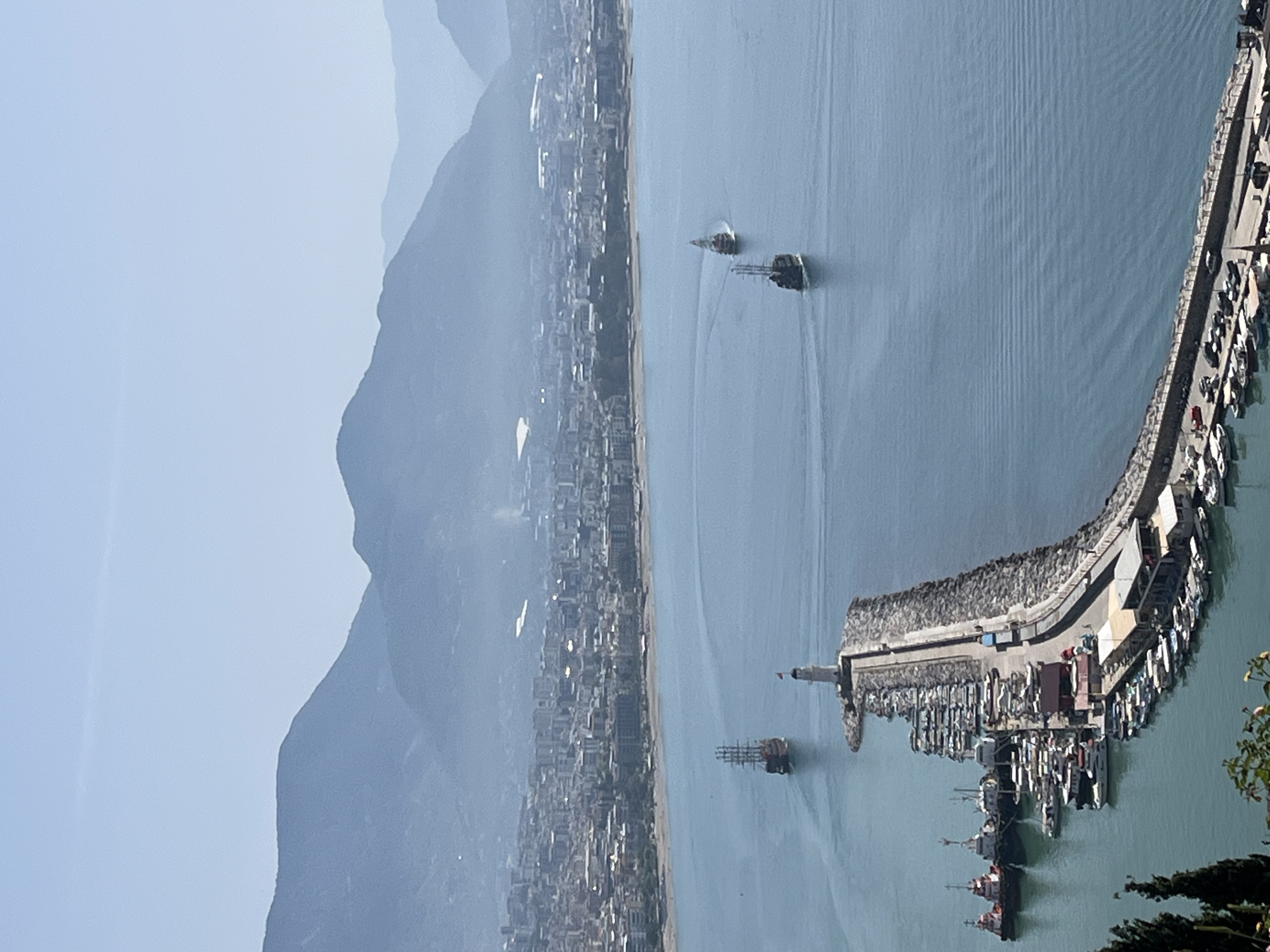The image shows a panoramic view of Alanya, Turkey, featuring mountains, a city skyline along the coastline, and the harbor. In the water, several boats are visible, including at least two distinctive tour boats with a pirate ship design. The harbor is characterized by a long, curved breakwater that shelters numerous docked vessels. A lighthouse is positioned at the end of the breakwater. The weather appears clear, with some haze obscuring distant features. Buildings dot the landscape near the coastline, marking the urban extent of Alanya. Mountains rise inland, creating a dramatic backdrop to the coastal city.