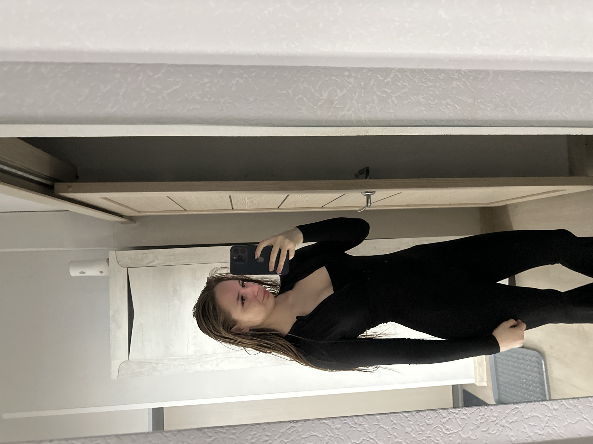 The image shows a woman taking a selfie in a mirror. She is wearing a black, form-fitting outfit with long sleeves. Her hair is wet and appears to be light brown. The background includes a bathroom vanity, a cabinet, and textured walls. The location is Unknown, Unknown.