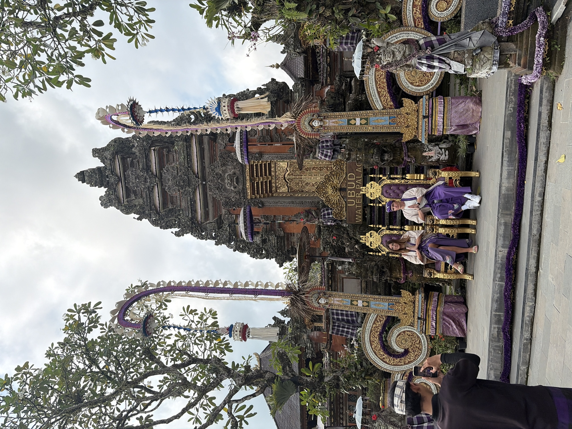 In front of the Ubud Water Palace in Ubud District, Indonesia, a man photographs a couple seated on ornate thrones. The couple, dressed in traditional Balinese attire, are positioned symmetrically on gold-accented thrones adorned with purple fabric. The man wears a white top and purple pants with a matching headband, while the woman is in a white top and a purple sarong. The palace facade, a multi-tiered stone structure with intricate carvings and golden doors, forms the backdrop. Decorative elements like large purple and gold floral motifs, traditional umbrellas, and statues of mythical figures flank the scene. A tree with lush green foliage extends from the left side of the frame, and the sky above is overcast. The man taking the photo, wearing a dark shirt and a traditional head covering, is captured mid-action, holding his phone horizontally to capture the couple. The ground in front of the thrones is paved, with a low step decorated with purple flower garlands leading up to them.