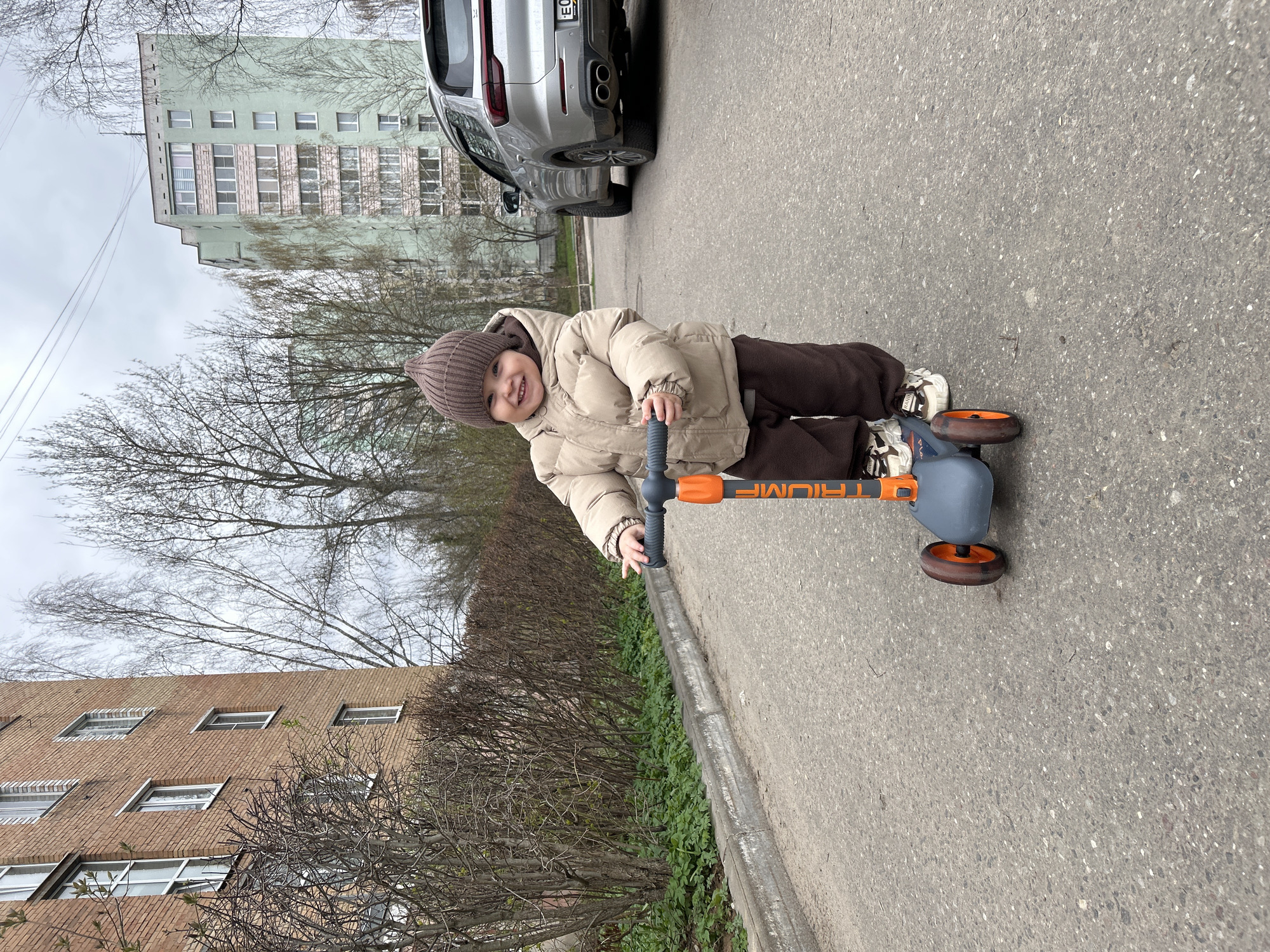 A child in a brown hat, beige jacket, and brown pants is riding a Triumf scooter on an asphalt road. The background includes a light green apartment building and a brick building with windows. A silver car is parked partially in the frame. Bare trees and shrubs are visible along the roadside. The setting appears to be in an urban area. The child is smiling. The car license plate is E093PX750.