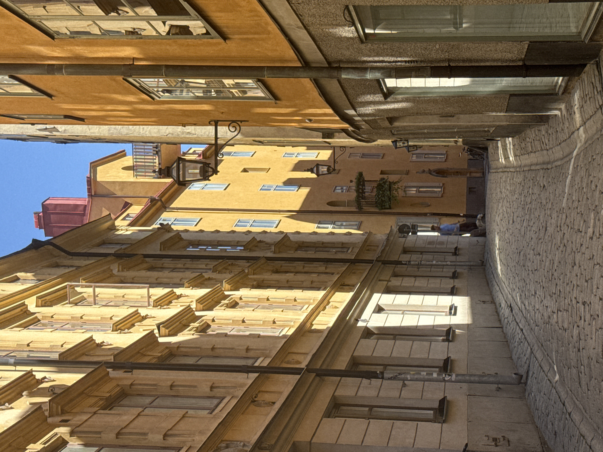 This image depicts a narrow, cobblestone street in Gamla Stan, Stockholm, Sweden. Tall, ochre-colored buildings with visible architectural details line both sides of the street. The buildings feature numerous windows, some of which are open, and ornate cornices and window surrounds. Several street lamps are mounted on the walls, casting shadows onto the pavement.
In the middle ground of the street, a single individual wearing a blue shirt and dark pants is walking away from the camera, towards a parked bicycle. The sunlight casts strong shadows across the cobblestone street, indicating a sunny day. The sky above is clear and blue. The overall impression is of a historic, well-preserved European streetscape.