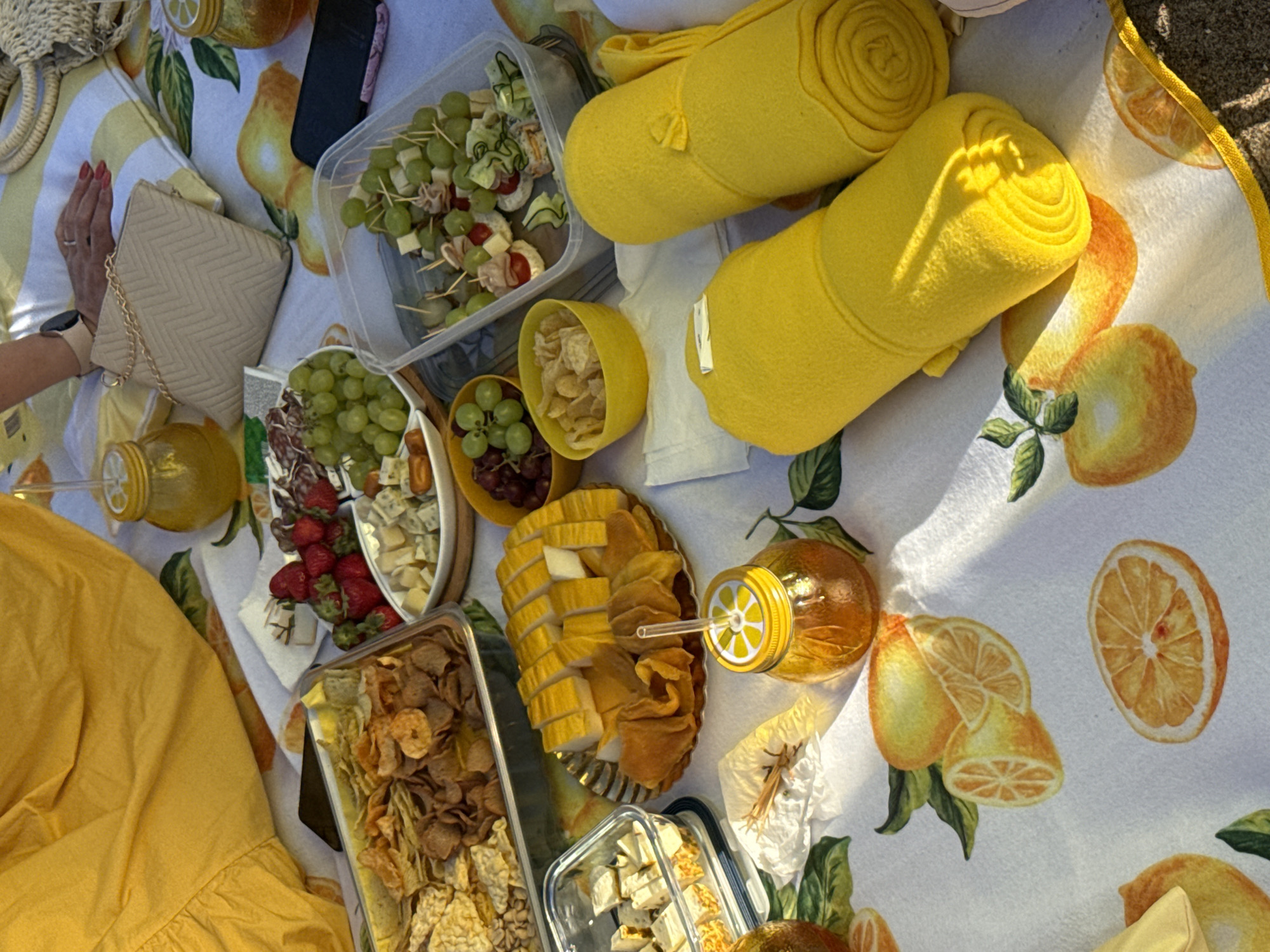 A high-angle, full shot captures a picnic blanket spread on a sandy surface, adorned with a vibrant lemon-themed print. The blanket is laden with various food items, including a tray of mixed chips and crackers, a platter of sliced cheese and salami, bowls of grapes and chips, and a dish of skewers with grapes, cheese, and cucumber. Two rolled yellow blankets lie on the right side of the frame, and a beige purse with a gold chain rests near a person's hand. A yellow-lid mason jar with a straw holds a golden liquid, and another similar jar is visible in the upper right corner. The lighting suggests it is daytime, with sunlight casting shadows across the scene. The overall atmosphere is one of a relaxed outdoor gathering.