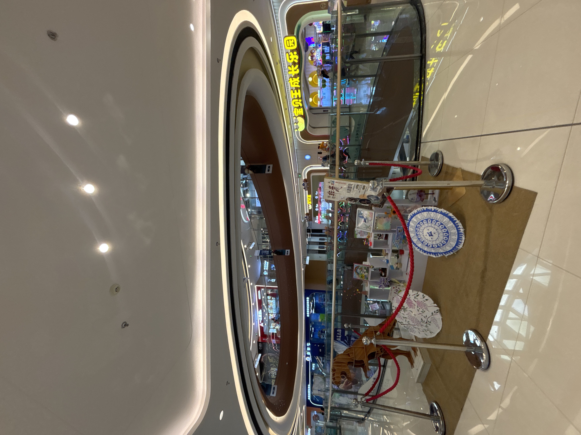 The image shows the interior of a modern shopping mall, likely in Jiangjin, China, featuring multiple levels and a contemporary architectural design. In the foreground, a display includes a wooden dinosaur model, two decorative umbrellas with intricate patterns, and several shelves filled with various items, possibly crafts or merchandise. Polished chrome stanchions with red ropes cordon off the display area. The floor is tiled with large, light-colored ceramic tiles that reflect the ambient light.
The background reveals the mall's multi-story structure with escalators and walkways. The upper levels are lined with shops, some illuminated with bright signage, including one with prominent yellow Chinese characters that translate to "Baby King Growth Paradise." The ceiling is a prominent feature, with a curved, multi-layered design and integrated LED lighting, creating a bright and airy atmosphere. Several circular light fixtures and what appear to be smoke detectors are visible on the ceiling. The overall scene depicts a commercial environment, likely a leisure or entertainment complex within the mall, with a focus on family-friendly attractions.