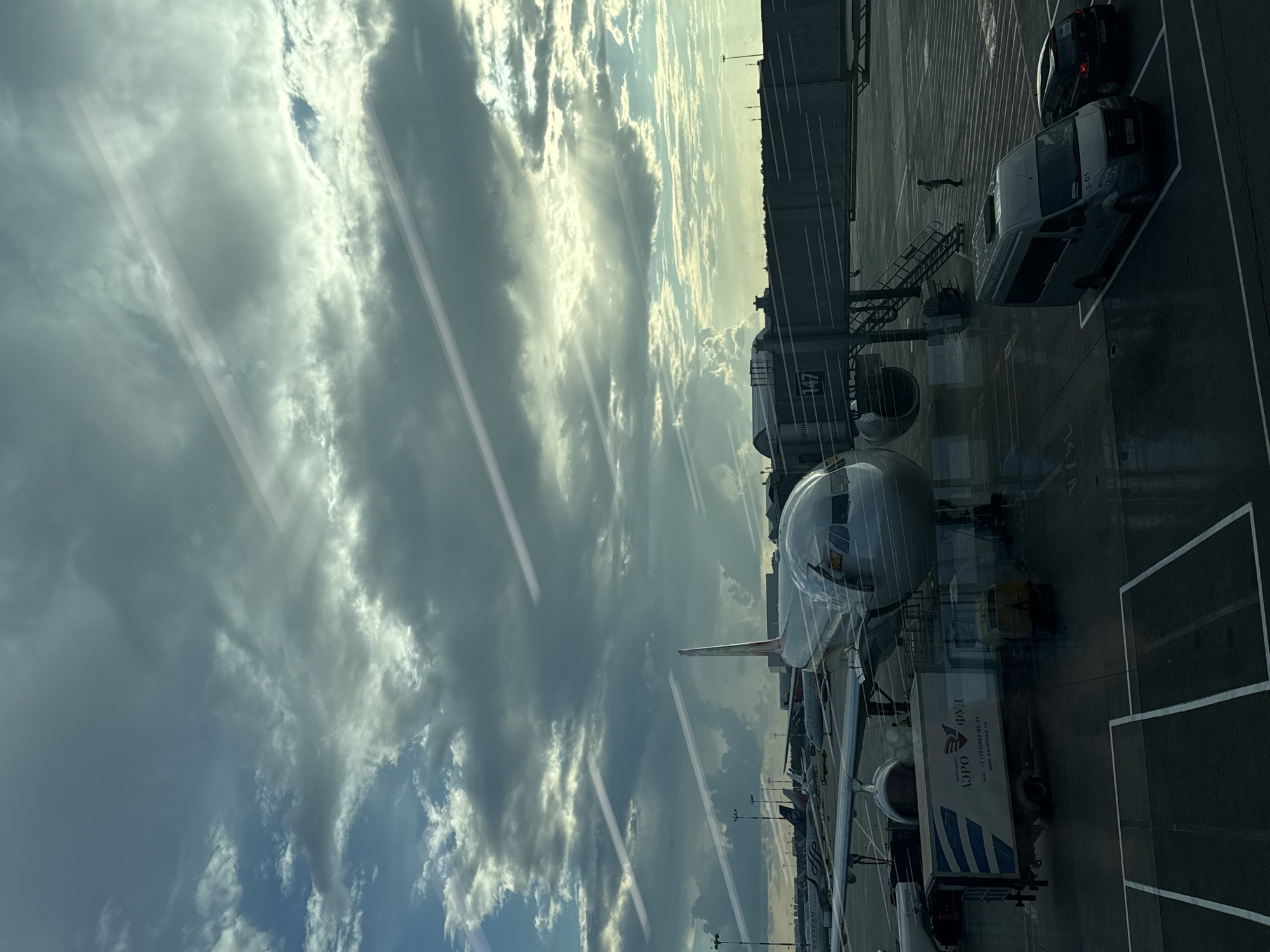 The image depicts an airport tarmac at the Sheremetyevo International Airport in Khimki, Russia, as seen through a window. A large passenger airplane is parked at a gate, connected by a jet bridge labeled with the number "147". Service vehicles, including a catering truck with "AERO FOOD" branding, are positioned around the aircraft. Several other aircraft are visible in the background, along with airport infrastructure like lights and terminals. The sky above is dramatic, with a mix of dark, heavy clouds and patches of sunlight breaking through, creating light rays. Two contrails are visible crisscrossing the sky. On the tarmac, a few individuals are present, including a person standing near the jet bridge stairs and another near a parked van. Two vehicles, a light-colored van and a dark car, are parked in designated spots. The ground appears wet, suggesting recent rain. The overall scene captures a moment at a busy international airport with interesting atmospheric conditions.
