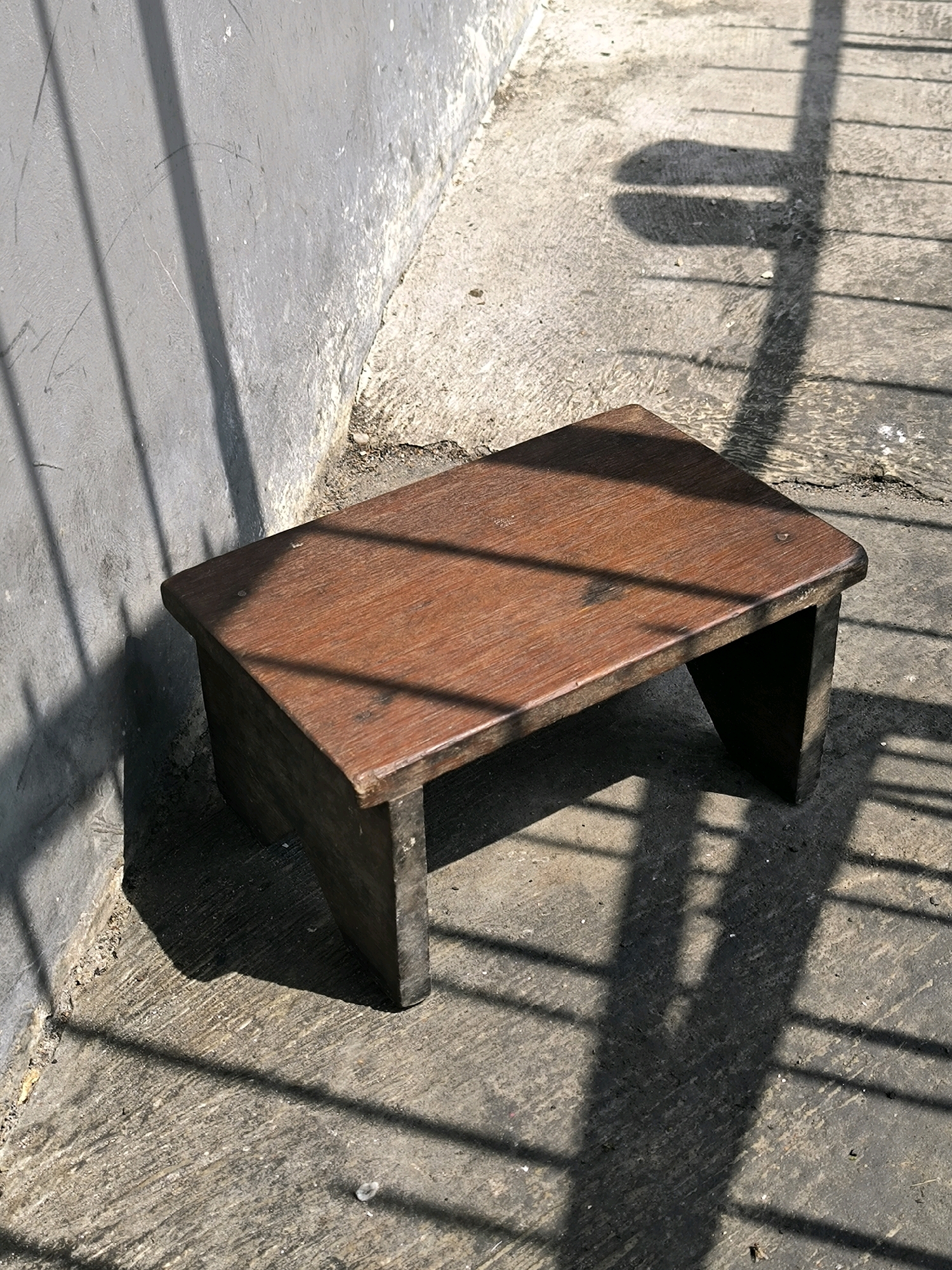 A low-angle, close-up, eye-level shot focuses on a small, dark wooden stool situated outdoors. The stool is positioned on a rough, grey concrete surface that appears to be a walkway or patio. A textured, light grey wall runs along the left side of the image, with shadows of vertical bars cast upon it. The concrete floor also bears similar linear shadows, suggesting the presence of a fence or similar structure overhead. The lighting indicates it is daytime, with strong sunlight creating distinct shadows. The stool has a simple, rectangular design with a dark wood finish and slightly rounded corners on its top surface. Its legs are short and appear to be made of the same wood. The scene is devoid of any people or animals. The overall impression is one of an ordinary, somewhat weathered outdoor space.