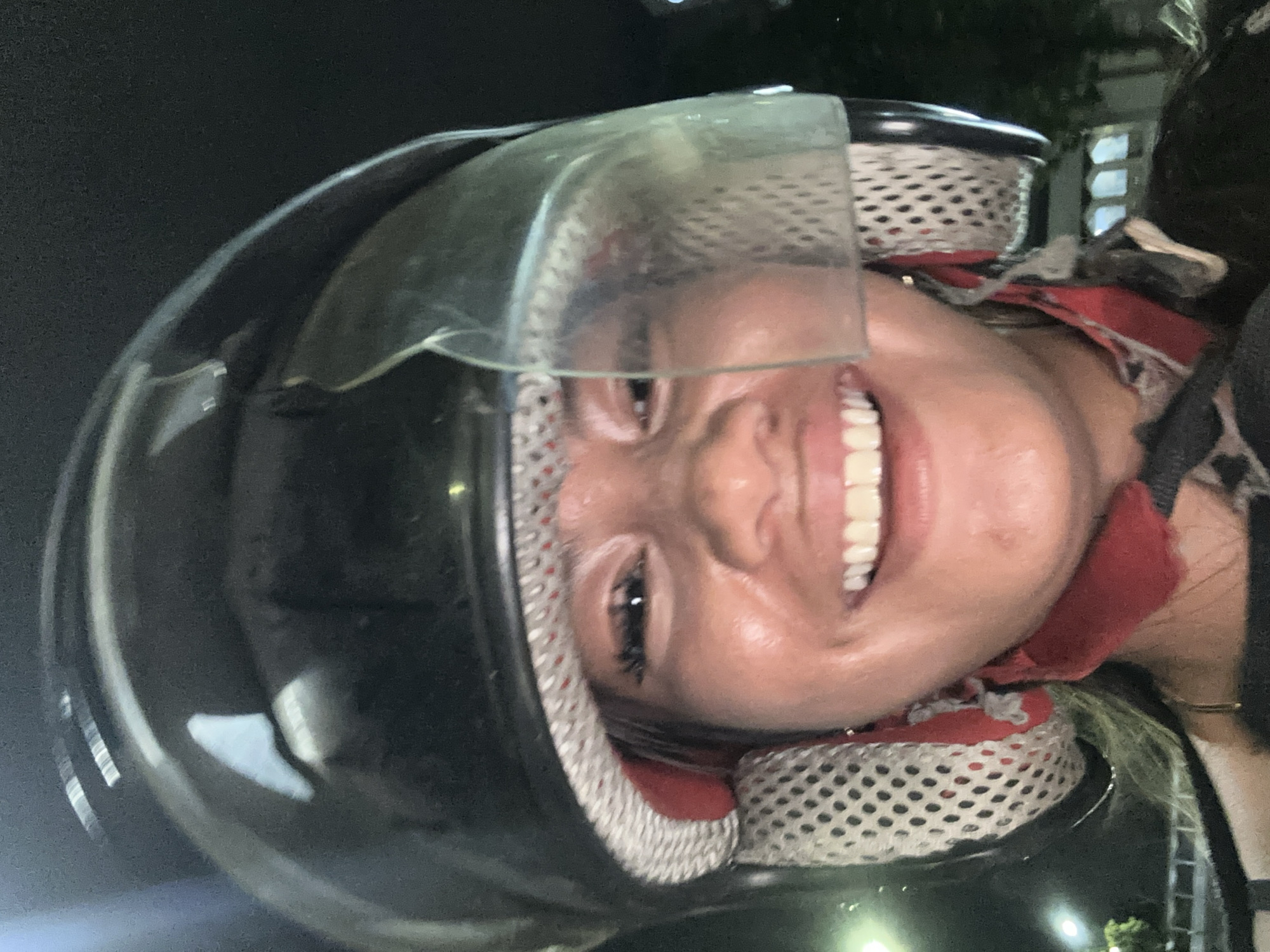 The image shows a woman wearing a black motorcycle helmet with the visor partially open. She is smiling. The helmet has a white and red padded interior. She is wearing a red and black garment under the helmet. The background is dark, with some indistinct light sources. Based on the location metadata (Central Jakarta, Indonesia), it's likely an urban street at night.