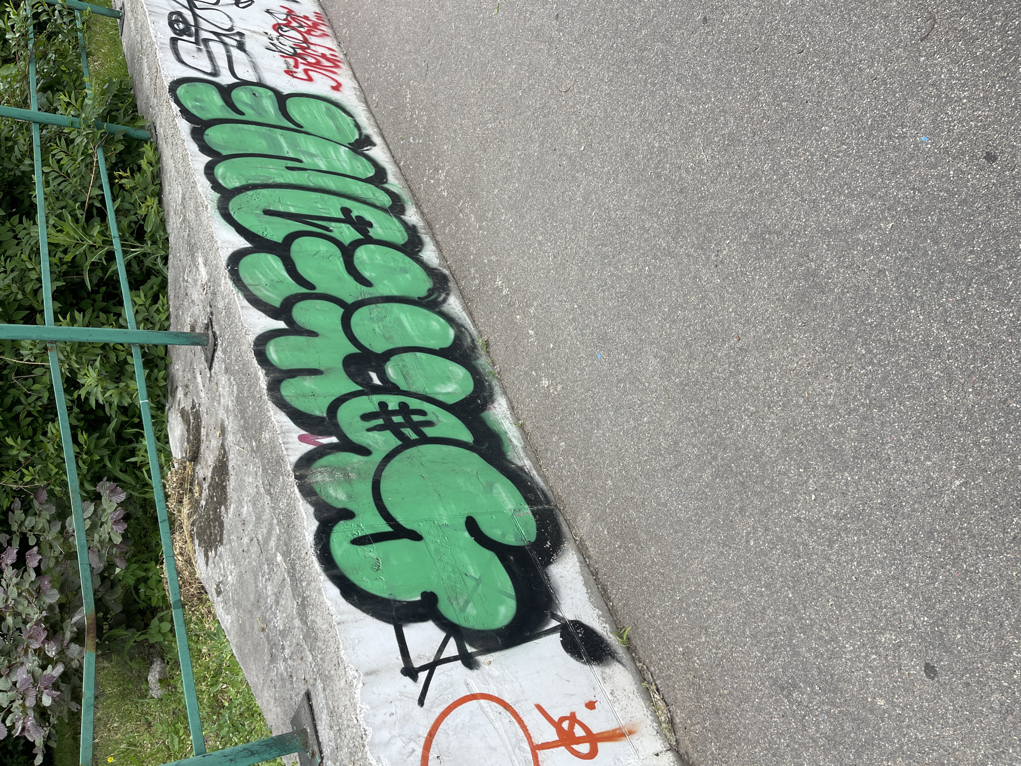 An outdoor, eye-level shot captures a concrete barrier running diagonally from the bottom left to the top right of the frame, separating an asphalt surface from an area of green foliage. The concrete barrier, which appears weathered and stained in places, is covered in graffiti.
The most prominent graffiti is a large, bubble-style piece in bright green with thick black outlines, spelling out "GREENS". The 'N' in "GREENS" is stylized, resembling a hash symbol (#). Above this green graffiti, a smaller, red tag reads "STR8 UP", and other indistinct black tags are also visible. Below the main green artwork, on the lower part of the concrete, there are additional orange graffiti elements, including a circular shape and various lines.
To the left of the concrete barrier, a green metal railing runs parallel to it, partially obscured by dense green bushes and foliage growing on the other side. Some dry weeds and dirt are visible at the base of the railing and concrete. To the right of the barrier is a gray asphalt surface, possibly a road or pathway, which appears mostly clear, with a few small specks and variations in its texture.
The scene is brightly lit by daylight, indicating a clear, sunny day. No people or activities are present.