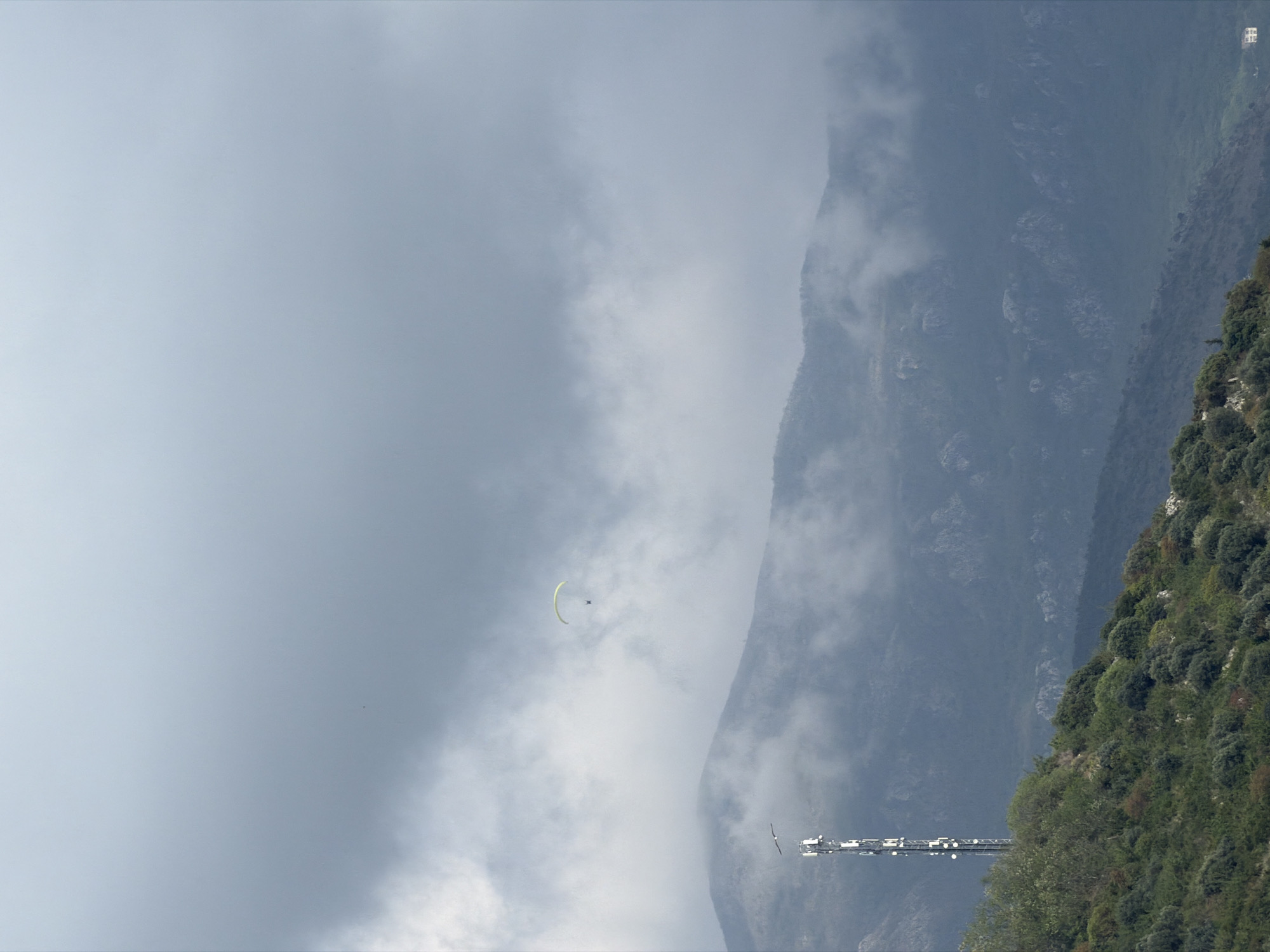 The image captures a vibrant paragliding scene against a dramatic mountainous backdrop in Budva, Montenegro. The setting features a rugged mountain range on the right side of the frame, its slopes densely covered with green vegetation, including trees and bushes. Clouds and mist partially shroud the mountains, creating a hazy, atmospheric effect.
In the open sky on the left, a paraglider with a bright, light green or yellow canopy is clearly visible, carrying a single individual. The paraglider appears to be soaring effortlessly, engaged in the activity of paragliding amidst the natural scenery.
Towards the lower right, at the base of the mountain, stands a modern communication tower. This structure features a tall, lattice design with several circular antenna dishes mounted along its height. A small bird can also be seen in flight near the tower. High on a distant peak in the upper right, a tiny white building or structure is faintly discernible.
The time of day appears to be daytime, with soft, diffused light indicating an overcast or cloudy weather condition. The overall mood is one of adventure and natural beauty, showcasing the potential for outdoor activities in a picturesque, mountainous coastal region. There is no visible text in the image.
