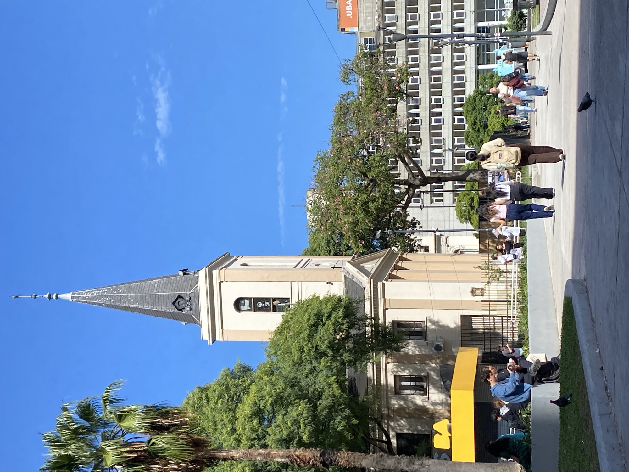 The image depicts an outdoor urban scene on a clear, sunny day in the Monserrat neighborhood of Buenos Aires, Argentina. The central structure is the Parroquia San Ignacio de Loyola church, located on the left side of the frame. It features a cream-colored facade, a multi-story bell tower with arched openings, and a dark, conical spire topped with a cross finial. Below and to the left of the church, a commercial building with a yellow awning, partially displaying "BA," is visible beneath a tall palm tree.
In the background, a large, multi-story grey concrete building with numerous horizontal rectangular windows serves as a prominent backdrop. An orange sign marked '.UBA' is visible on the rooftop of a building further to the right in the distance, identifying a University of Buenos Aires location.
The foreground consists of a grey paved pedestrian pathway bordered by concrete planters containing green grass. On the left, a group of approximately 5-7 individuals are seated on a concrete planter. One male, wearing a blue hoodie and dark pants, is holding a mate gourd and a thermos. Other people are seen walking along the paved path; in the midground, three women walk away from the viewer on the right, one carrying a light brown backpack. Additional pedestrians are present further in the background. Two pigeons are observed walking on the paved ground, one near the seated group and another on the right side of the path. Green deciduous trees with some pink blossoms are dispersed throughout the scene, all under a bright blue sky with sparse white clouds.