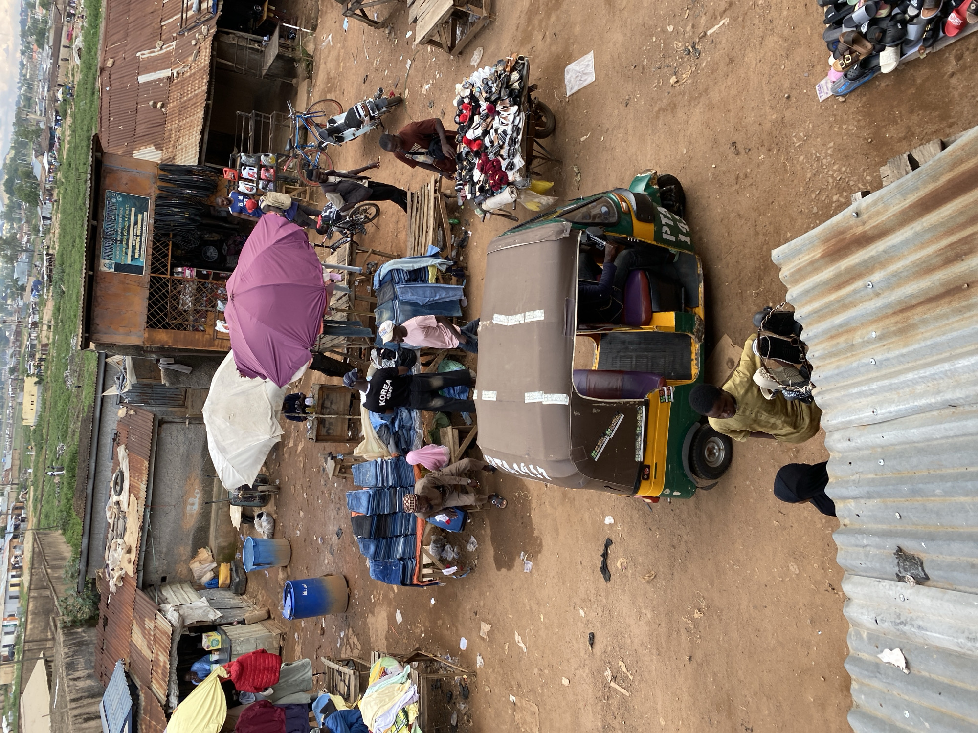This image depicts an active, open-air market scene in Bukuru, Nigeria, viewed from a high vantage point looking down onto a dirt ground. The environment is characterized by makeshift stalls with corrugated metal roofs, some rusted, and a general impression of an unpaved, utilitarian commercial area. In the mid-ground, a central clearing of reddish-brown dirt is surrounded by various vendors and their goods. To the left, a stall with a blue corrugated roof and a yellow awning is partially visible, showing a man in a blue shirt behind a counter. Adjacent to this, two large blue plastic barrels are on the ground. Further right, a white umbrella shelters a vendor, whose presence is partially obscured. Towards the center, a large purple umbrella provides shade over a display area. Behind this, a building with a rusted corrugated roof features a prominent sign reading "CHINOYS O. ABIC INVESTMENT," indicating a business selling tires, motor oil, and other automotive parts, displayed outside. Two men are standing near this shop. Two motorcycles and two bicycles are parked nearby. In the center of the dirt area, goods are displayed on low wooden tables. A significant pile of folded blue jeans is stacked high on one table, with a man in a brown traditional cap and attire seated beside it, along with a child in a pink hijab. Another stack of jeans is visible to the right, where a man in a black "KOREA ARMY" t-shirt stands. Further right, a man in a brown t-shirt tends to a pushcart overflowing with numerous pairs of shoes and sandals. A prominent green and yellow three-wheeled motorized rickshaw (tricycle), marked "PTA 14..." on its side, is positioned in the mid-ground, facing away from the viewer. Its driver is partially visible inside. In the foreground, the edge of a galvanized corrugated metal roof is visible. Below it, two individuals are seen from behind: one wearing a black head covering and another in a light brown long-sleeved shirt, carrying a bag. The ground throughout the market is unpaved, uneven, and covered in reddish-brown dirt, with scattered debris including plastic bags and other refuse. In the distant background, beyond the market stalls, residential or commercial buildings are visible, interspersed with green areas and what appears to be a more developed urban landscape under a cloudy sky. At least twelve individuals are discernible within the scene, engaged in various market activities such as