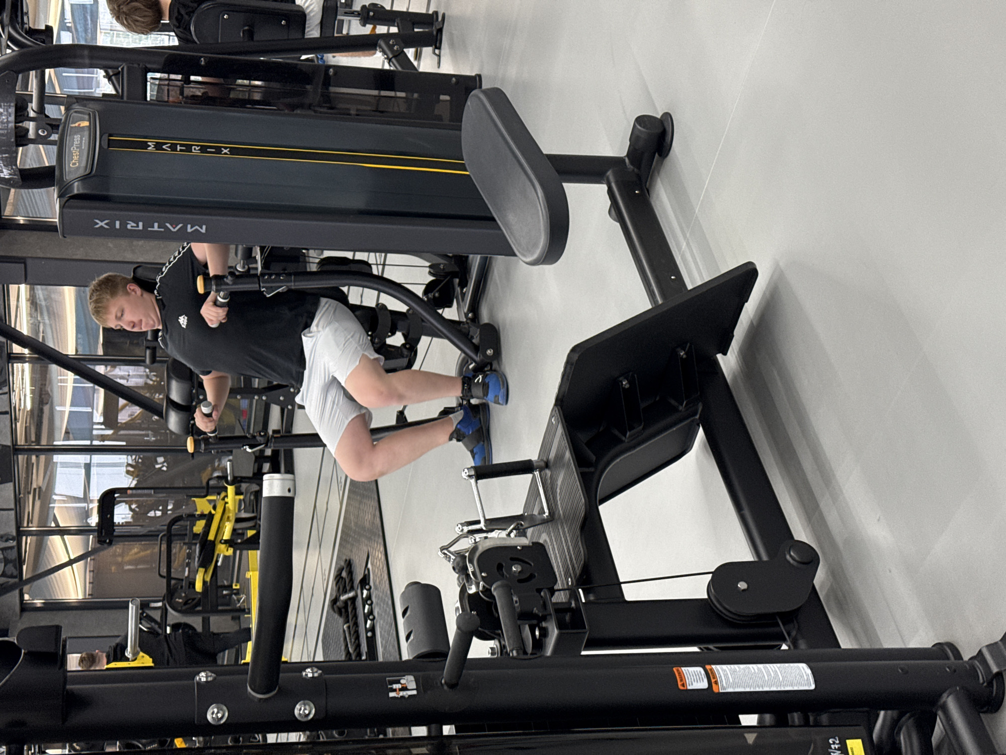 The image depicts the interior of a gymnasium. A young man is using a chest press machine from the manufacturer Matrix. The man is wearing a black t-shirt with white stripes on the sleeves and light gray shorts. He is wearing blue sneakers. Other exercise machines can be seen in the background, some of which are yellow. The floor is light gray. It is not possible to tell the exact location of the image.