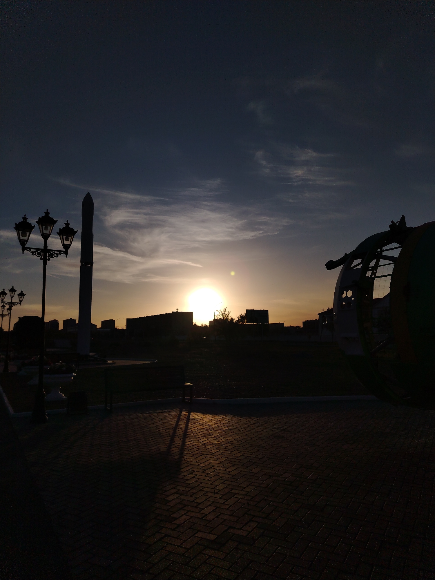 The image captures a silhouetted scene at sunset in Baikonur, Kazakhstan. The sun, a bright orb low in the sky, casts a warm glow, illuminating thin, wispy clouds against a darkening blue sky. In the foreground, a brick-paved walkway is shadowed by a decorative street lamp with three globes and a tall, slender monument that appears to be a rocket. To the right, a large, rounded structure, possibly part of an exhibit or a dismantled aircraft, is also silhouetted. The background features a line of buildings and trees, all rendered in dark silhouette against the fading light. The scene evokes a sense of quiet contemplation, with the setting sun and long shadows suggesting the end of the day. The location context points to a place of significance related to space exploration, given Baikonur's history as a cosmodrome. No visible text is present in the image.
