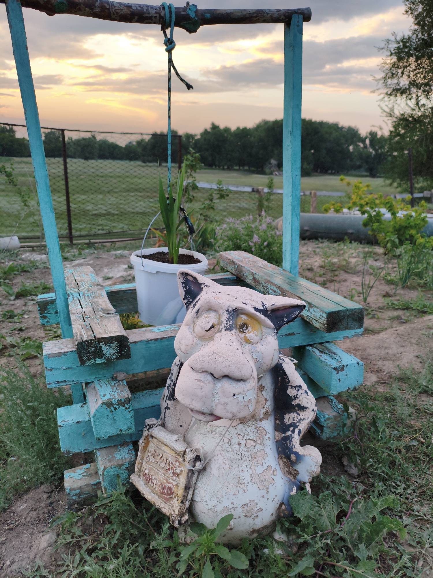 The image depicts a weathered garden gnome-like statue positioned in front of a rustic wooden structure painted teal, resembling an old well. The statue, made of a white material with peeling paint, has a stylized animal face and holds a small, framed object. Behind the structure, a fence encloses a grassy field with trees in the distance. The sky above shows a sunset with soft clouds. The scene is set outdoors, with overgrown grass and dirt on the ground. There are no people visible. The location is identified as Ақсуат ауылдық округі, Kazakhstan.