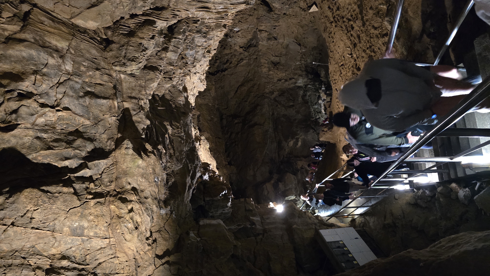 The image shows a group of people descending a metal staircase inside a cave. The cave walls are rough and textured, with visible layers of rock. The staircase is well-lit, creating highlights on the metal railings and the faces of the people.
The subjects are a mix of men, women, and children, all dressed in casual clothing. Some are wearing hooded jackets. They appear to be on a guided tour or exploring the cave. The activity is walking down the stairs, and the overall mood seems neutral, with no strong emotions evident.
Notable details include the texture of the cave walls, which vary from smooth to jagged. The lighting is artificial, casting shadows that emphasize the cave's natural formations. There is a white panel with some text on it visible at the bottom left of the image, but the text is not legible.
The scene is set within a cave, likely a tourist attraction given the presence of a sturdy staircase and railings. The location context provided is Vavřinec, Czechia, suggesting this is a cave in that region. The time of day and weather are not discernible from the image.