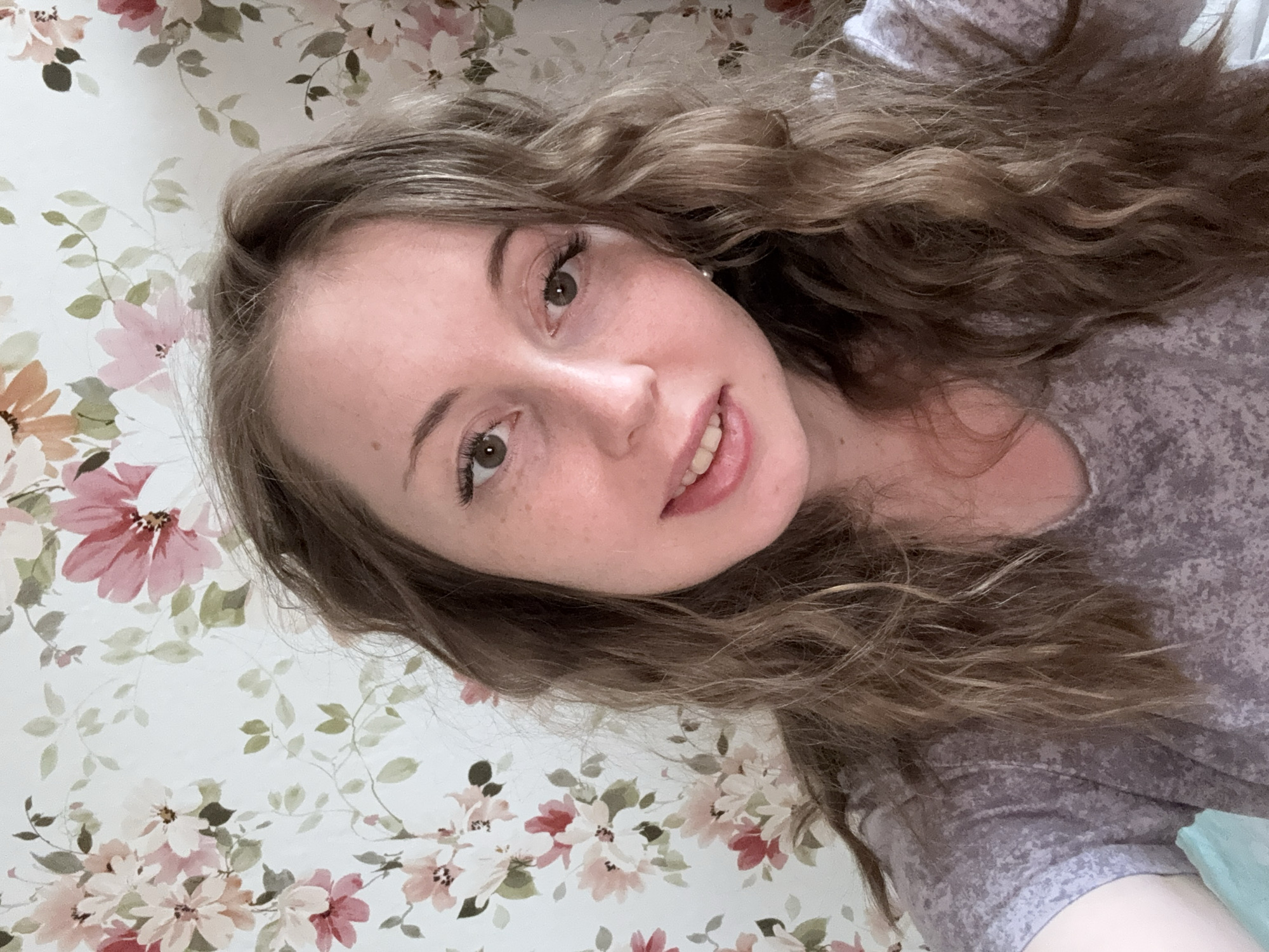 A young woman with curly light brown hair, freckles, and light eyes is photographed indoors against a floral wallpaper. She is wearing a grey t-shirt. The city and exact location are not discernible from the image.