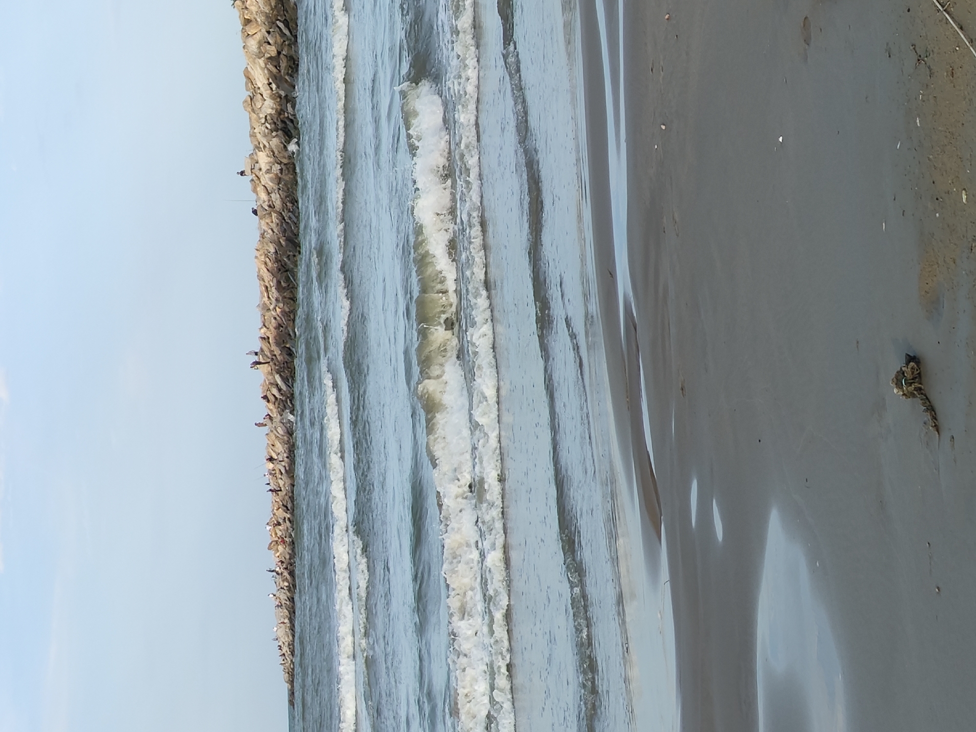 This image depicts a beach scene, likely in Makhachkala, Russia, featuring a breakwater made of large rocks extending into the sea. Numerous individuals are visible standing and fishing along the top of the breakwater. The sea is calm with gentle waves rolling towards the shore, creating white foam as they break. The foreground shows a sandy beach with some tidal pools and a piece of rope lying on the sand. The sky is partly cloudy.