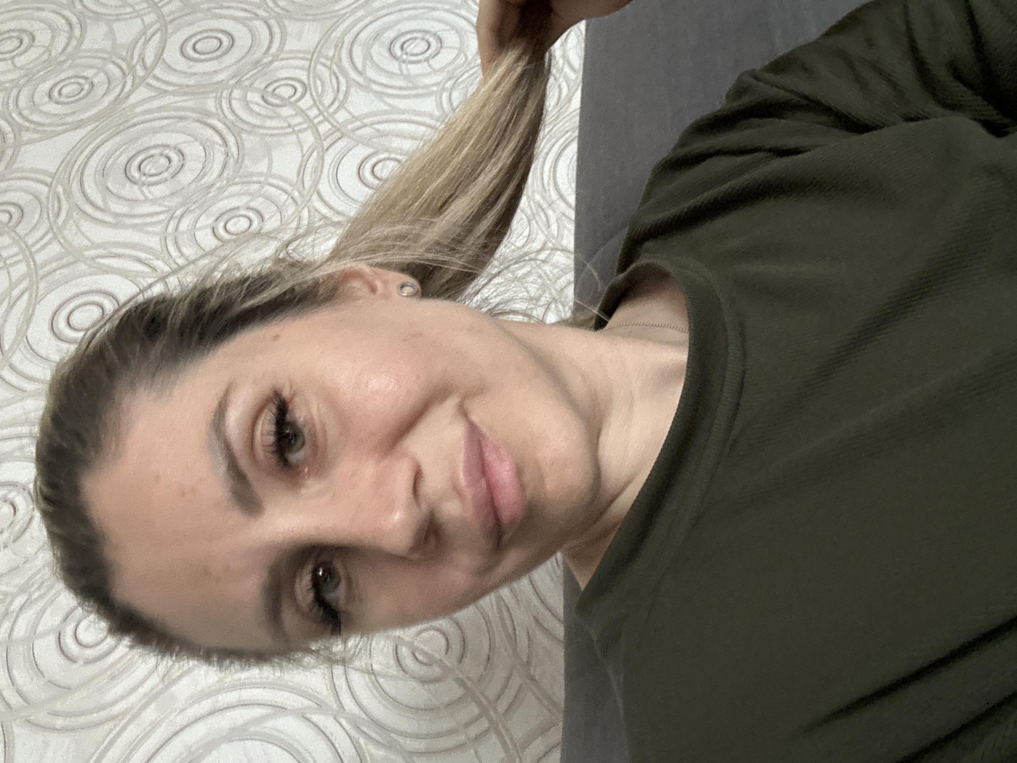 This is a close-up, selfie-style photograph taken indoors. The subject is a woman with blonde hair pulled back into a ponytail, and she is wearing an olive green long-sleeved shirt. She is looking towards the camera, and her left hand is touching her hair. The background consists of patterned wallpaper with concentric circles. The image provides no information about the specific location within Novoaltaysk, Russia.