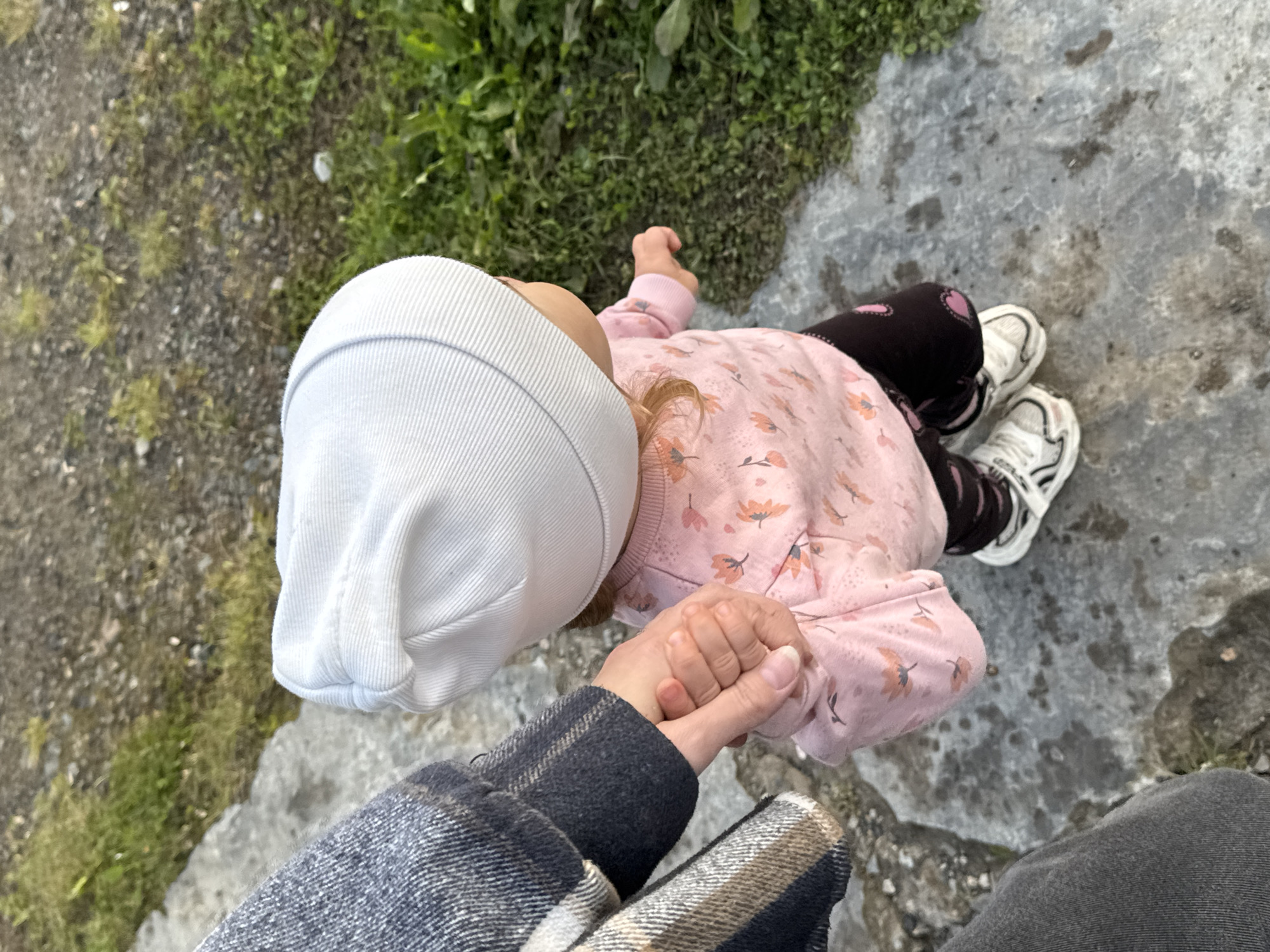 A child wearing a white beanie, pink long-sleeved shirt with a floral pattern, dark pants with a heart pattern, and white sneakers is walking on a stone path outdoors. The child's hand is being held by an adult wearing a plaid jacket. The environment is rural, with grass and dirt visible in the background. The image appears to have been taken in Комарихинский, Russia.
