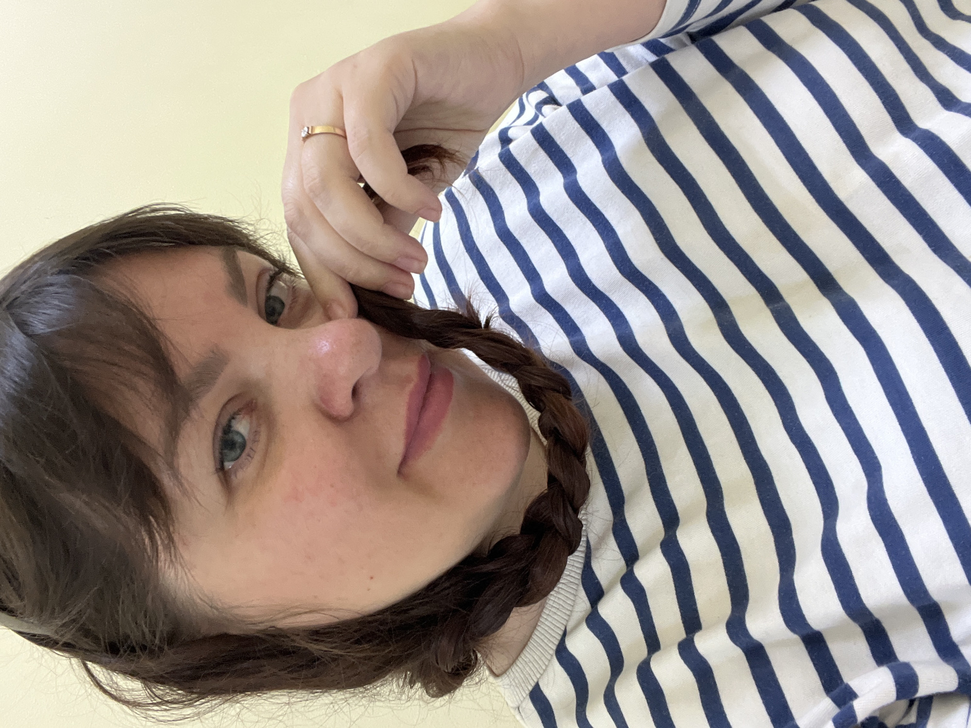 A woman with brown hair in two braids and bangs, wearing a blue and white striped shirt, is seen in a close-up, selfie-style shot. She has light skin and blue eyes and is touching her face with her right hand. A gold ring with a small stone is visible on her right ring finger. The background is a plain, light yellow wall. The image does not contain any information that would allow for the determination of its location in Noviy, Russia.