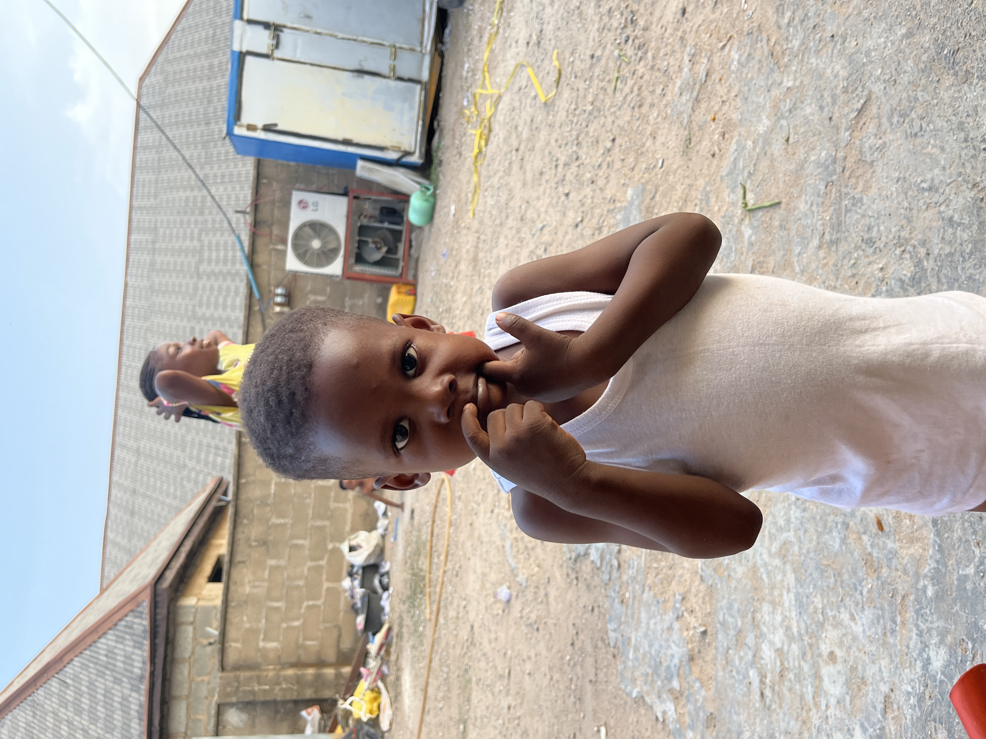 A young, dark-skinned child with close-cropped hair stands in a yard. They are wearing a white sleeveless shirt and have their fingers touching their face. In the background, there are buildings constructed of gray concrete blocks, an air conditioning unit, and various pieces of trash and debris scattered across the yard. Another person wearing a yellow shirt can be seen standing in the background behind the child. The setting appears to be a residential or commercial area in Gwagwalada, Nigeria.
