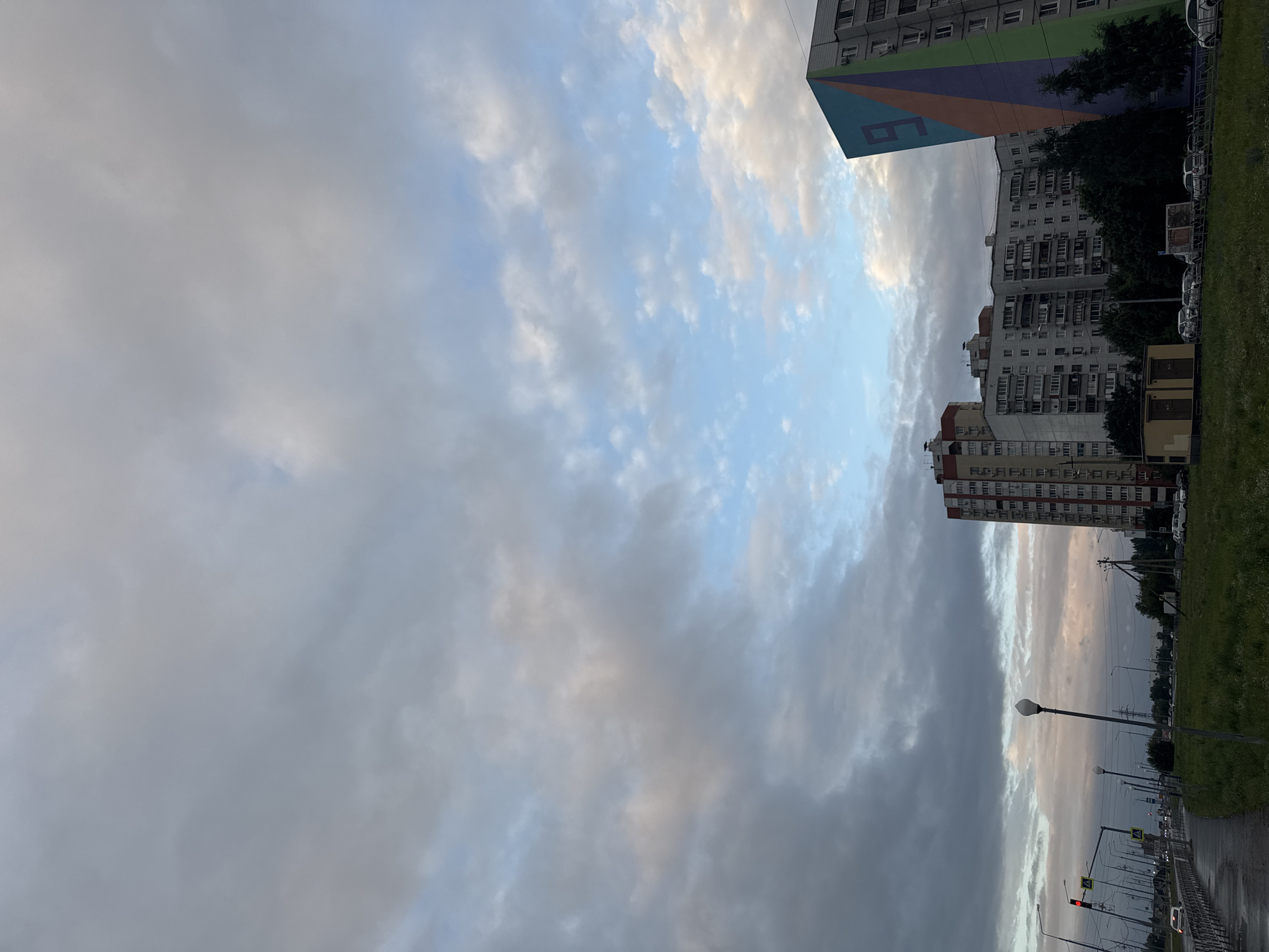 The image depicts a street scene in Tolyatti, Russia, under a cloudy sky. On the right, a tall residential building is visible, notable for a large, colorful geometric mural on its side, featuring shades of blue, purple, orange, and green, with the numeral "9" prominently displayed. Several other apartment buildings, with multiple floors and windows, are situated behind the muraled structure, forming a typical urban residential landscape. In the foreground, a patch of green grass with scattered white wildflowers borders a paved area, likely a sidewalk, which is wet, suggesting recent rain. A streetlamp with a white spherical light fixture stands on the sidewalk. To the left, a road with multiple lanes is lined with traffic lights, streetlights, and various vehicles, including cars and what appears to be a truck. Pedestrian crossings are marked. The overall atmosphere is overcast and somewhat somber, typical of cloudy weather.
