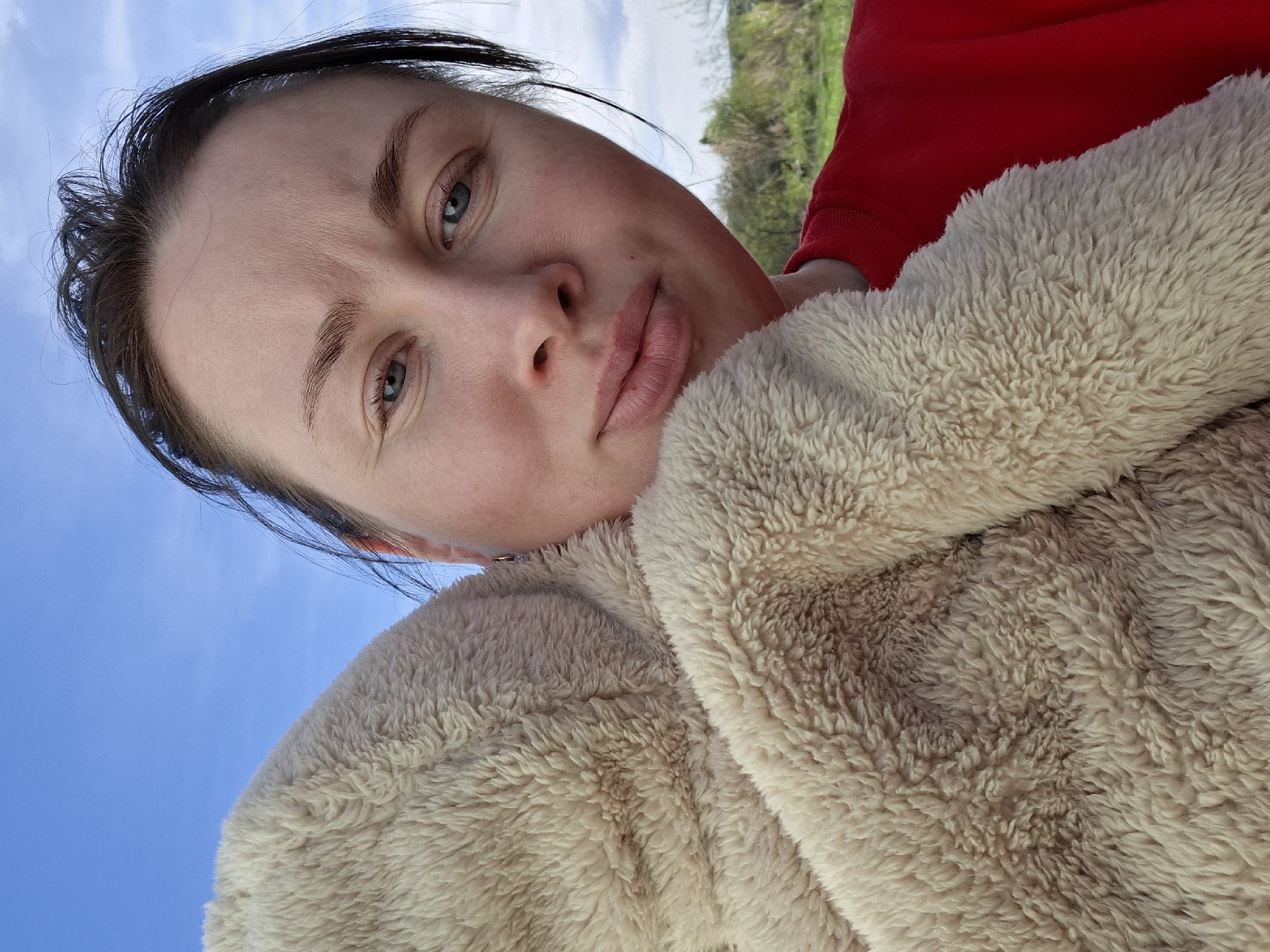 A close-up, tilted, selfie-style shot features a woman with fair skin, blue eyes, and dark hair pulled back. Her expression is neutral. She is wearing a fluffy, light beige garment, possibly a coat or jacket. A glimpse of a red garment is visible behind the fluffy material. The background consists of a blue sky and some greenery. The location is Unknown, Unknown. There are no discernible actions or interactions beyond the woman taking a selfie.