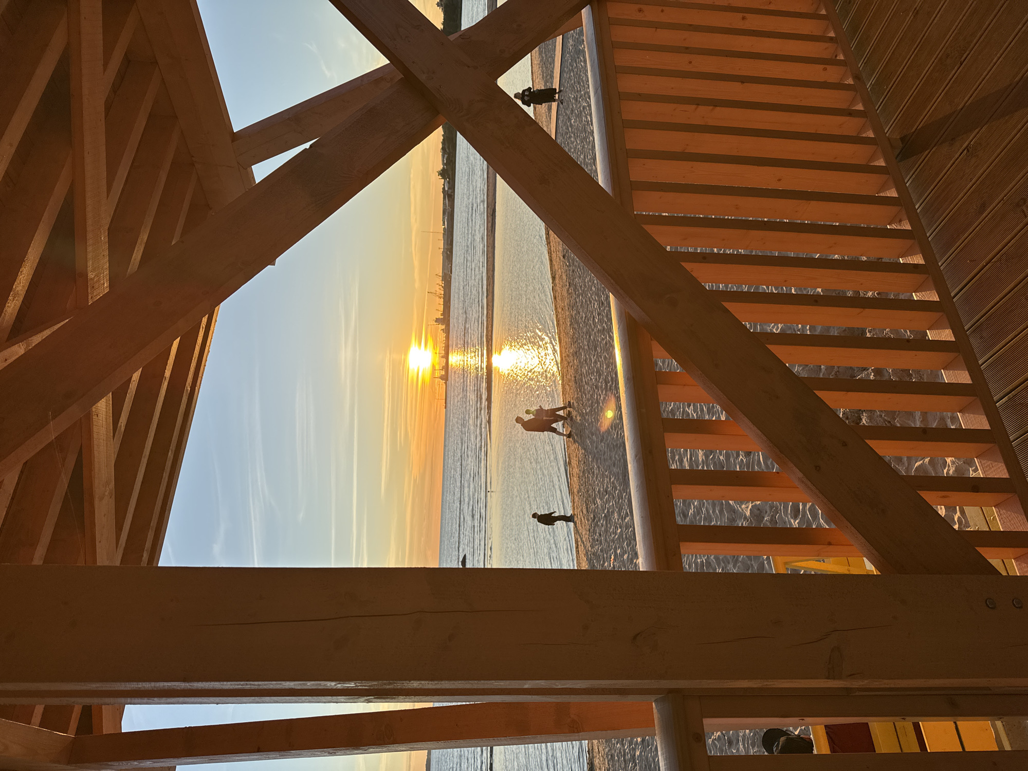 The image captures a sunset scene on a beach in Tallinn, Estonia, viewed from a wooden structure with a railing and overhead beams. The sun is low on the horizon, casting a golden glow on the water and sand. Several people are visible on the beach, including a group of three walking together and a solitary figure standing by the water. In the distance, industrial cranes and structures are silhouetted against the sky, indicating a port area. The wooden platform in the foreground is bathed in warm light, with shadows cast by the railing and structural elements. The sea is calm, reflecting the colors of the sunset.