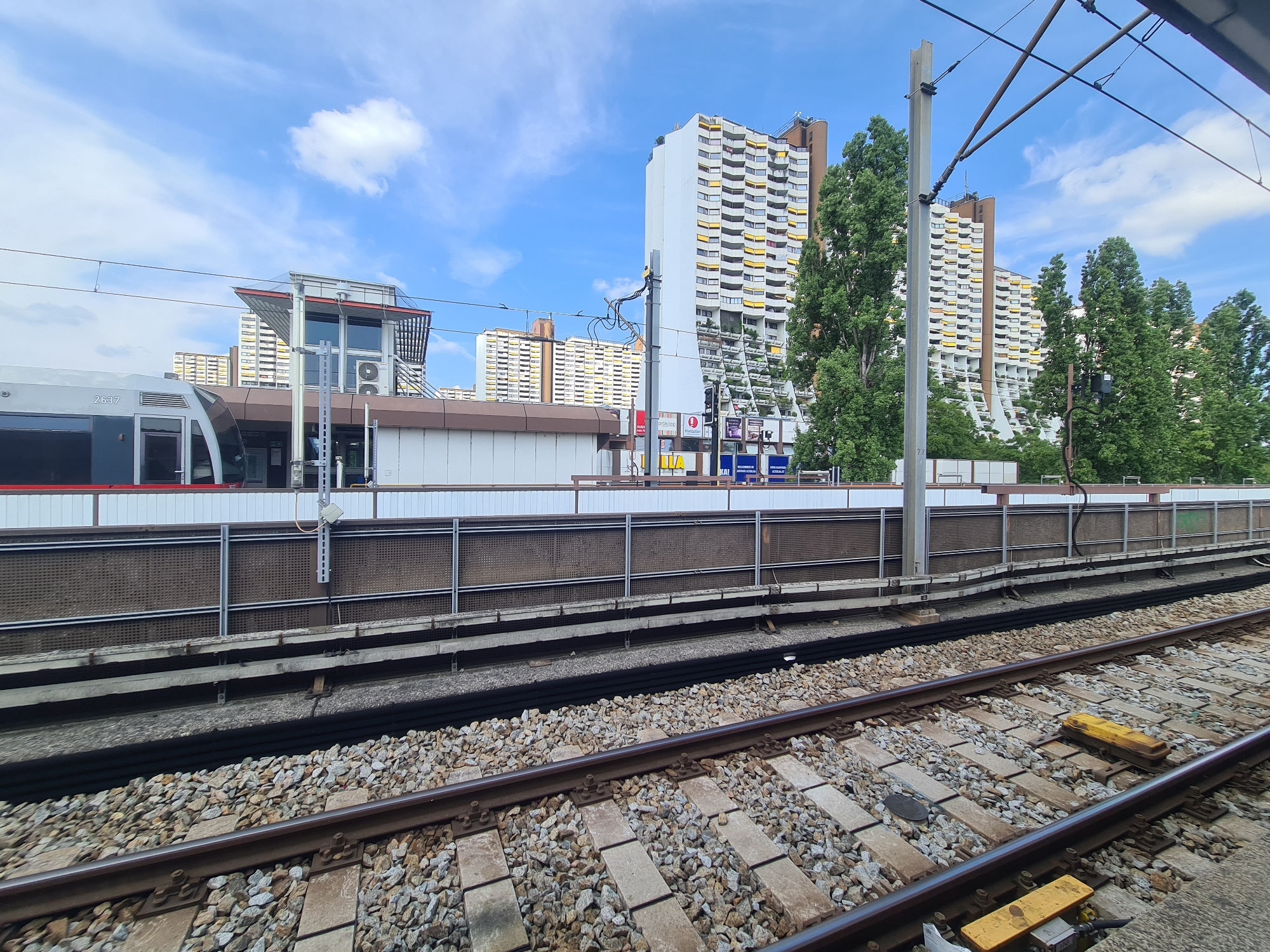 The image captures a scene at a train station in Vienna, Austria, under a bright blue sky with scattered clouds. In the foreground, a set of train tracks laid on gravel ballast is visible, with the rails running diagonally across the frame. Two yellow warning markers are present on the tracks in the lower right quadrant. Beyond the tracks, a platform is present, separated by a metal fence with a brown mesh infill. A train, identified by the number "2637" on its side, is stopped at the platform.
Behind the platform, a low-rise building with a brown trim is visible, possibly part of the station infrastructure. Further in the background, two towering apartment buildings with a distinctive architectural style, featuring white facades and numerous balconies with yellow awnings, dominate the skyline. These buildings are characteristic of the "Hundertwasserhaus" or similar designs, suggesting a location in the vicinity of such architecture. Lush green trees partially obscure the lower sections of these apartment blocks. Overhead, a complex network of power lines and support structures for an electric railway system stretches across the scene. The overall impression is one of a functional urban transit environment within a visually striking residential area of Vienna.