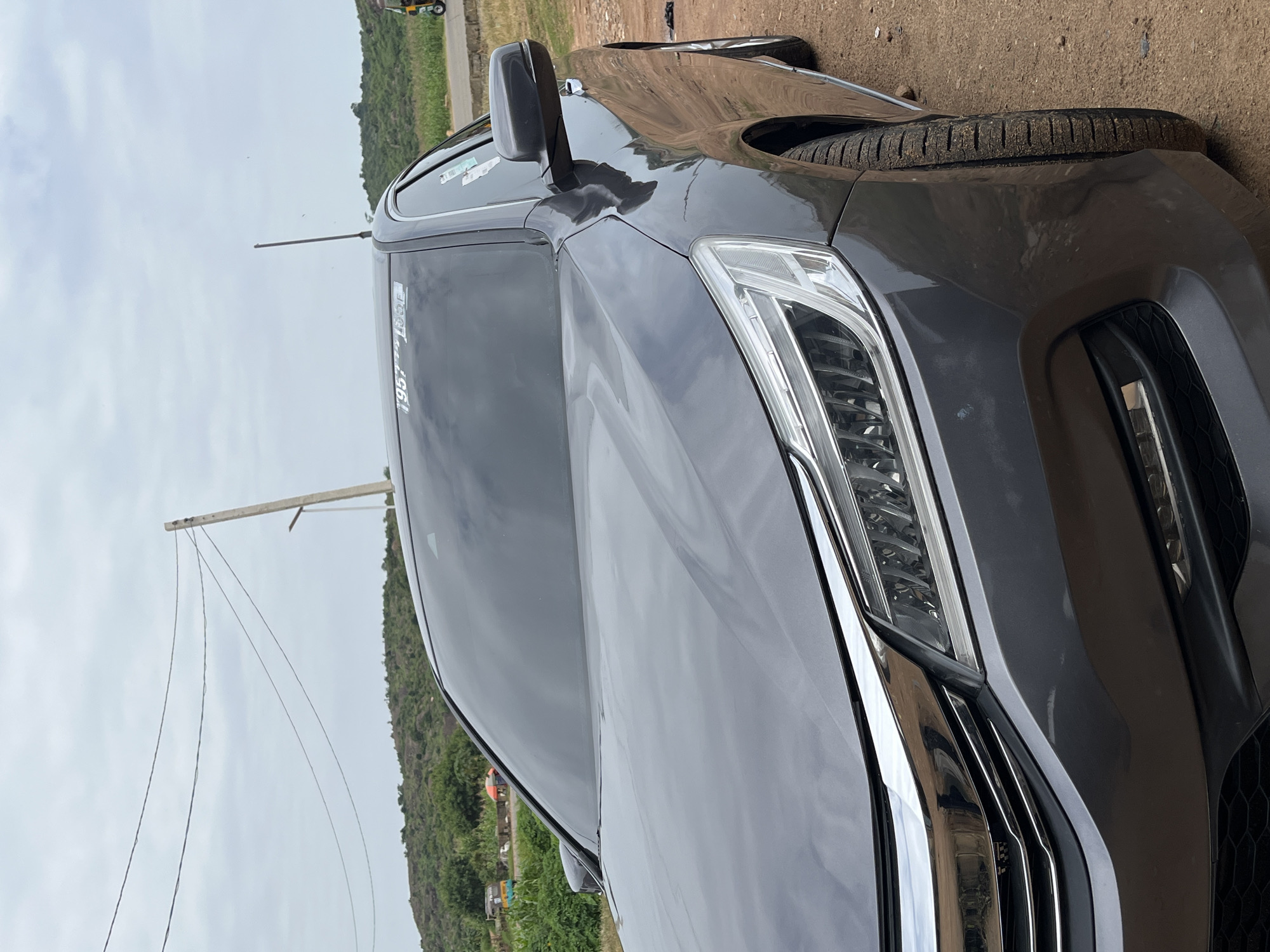 The image displays a close-up, low-angle view of the front of a dark gray sedan parked on a dirt road. The car's headlights and grille are prominent, with chrome accents visible. The windshield is tinted, obscuring the interior. In the background, a lush green, hilly landscape is visible under an overcast sky. Power lines stretch across the upper portion of the frame, supported by a utility pole. A small, colorful three-wheeled vehicle, possibly a Bajaj Auto, is parked in the distance on the right side of the frame. The overall impression is of a car parked in a semi-rural or developing area, likely on the outskirts of a city. The specific location in Jos, Nigeria, cannot be definitively determined from the visual cues alone, but the presence of a Bajaj Auto and the characteristic landscape are consistent with such a setting.