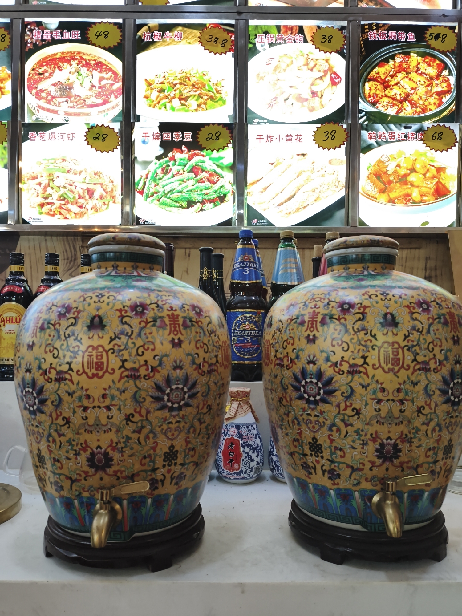 The image captures a close-up view of two large, ornate ceramic dispensers, likely for beverages, situated on a counter in what appears to be a restaurant or food establishment. These dispensers are decorated with intricate patterns featuring floral motifs and Chinese characters, primarily in yellow, blue, red, and green. Each dispenser has a golden spigot at its base and rests on a dark wooden stand.
In the background, a menu board displays images of various Chinese dishes with their prices. To the left and right of the dispensers, several bottles are visible, including some with labels that suggest alcoholic beverages like coffee liqueur and beer. The lighting in the background is bright, illuminating the menu items.
The scene suggests a setting where customers can serve themselves drinks from the dispensers while perusing the food options. The overall impression is one of a functional yet decorative establishment, possibly a casual eatery in Jixi, China, given the presence of Chinese script on the menu and the style of the food displayed. There are no people visible in the image.