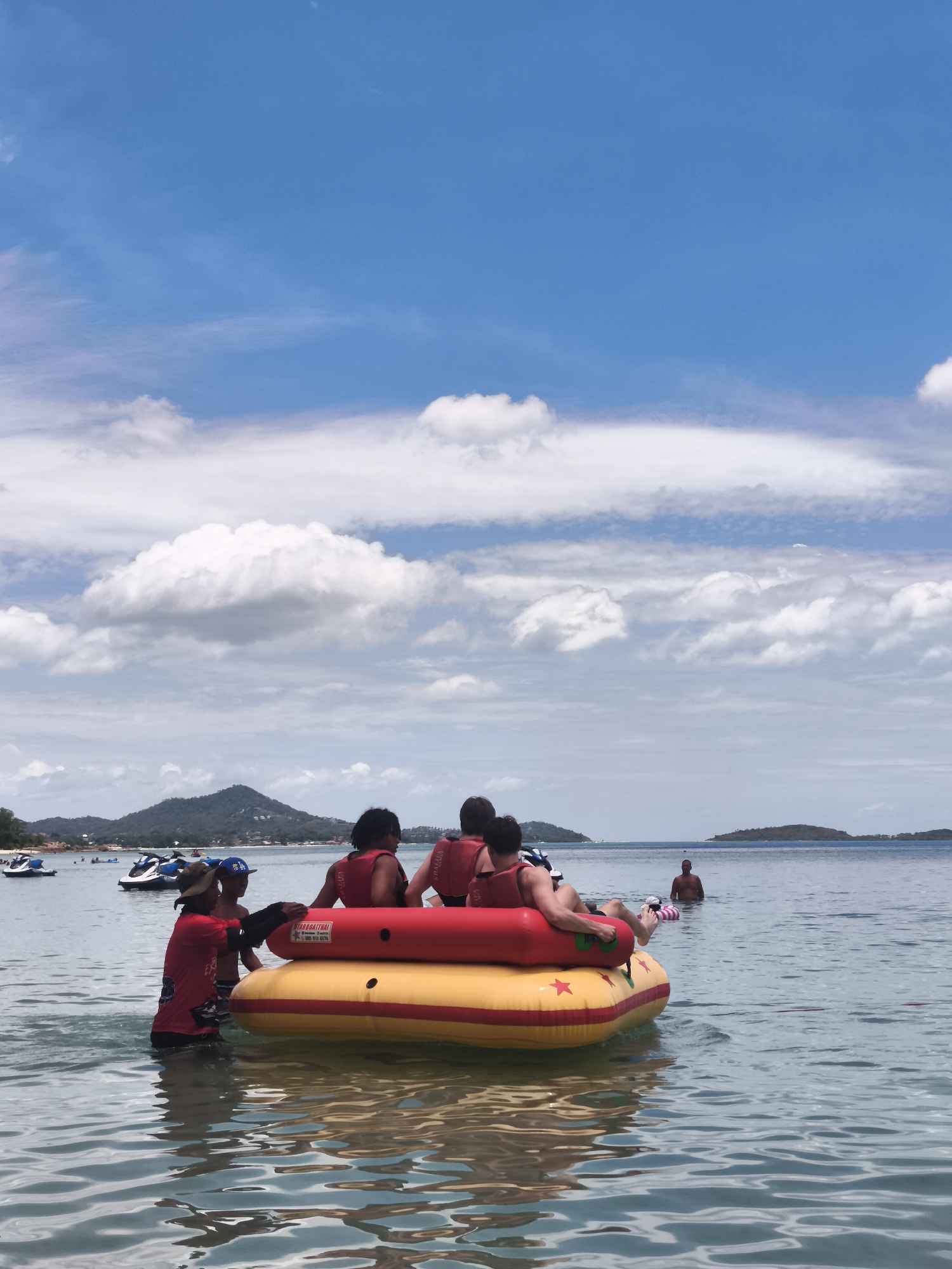 The image captures a bright, sunny day at a beach in Ko Samui District, Thailand. In the foreground, a yellow and red inflatable raft, possibly a banana boat, is partially submerged in the clear, shallow water. Four individuals are seated on the raft, all wearing life jackets, with their backs to the camera. A person in a red shirt and a hat, likely an attendant, stands in the water to the left of the raft, interacting with two younger individuals on the raft. In the mid-ground, several jet skis are visible near the shore, along with other people enjoying the water. The background features a lush, green island with scattered buildings, and the vast blue sky is adorned with scattered white clouds. The overall scene depicts a leisurely day of water activities in a tropical setting.