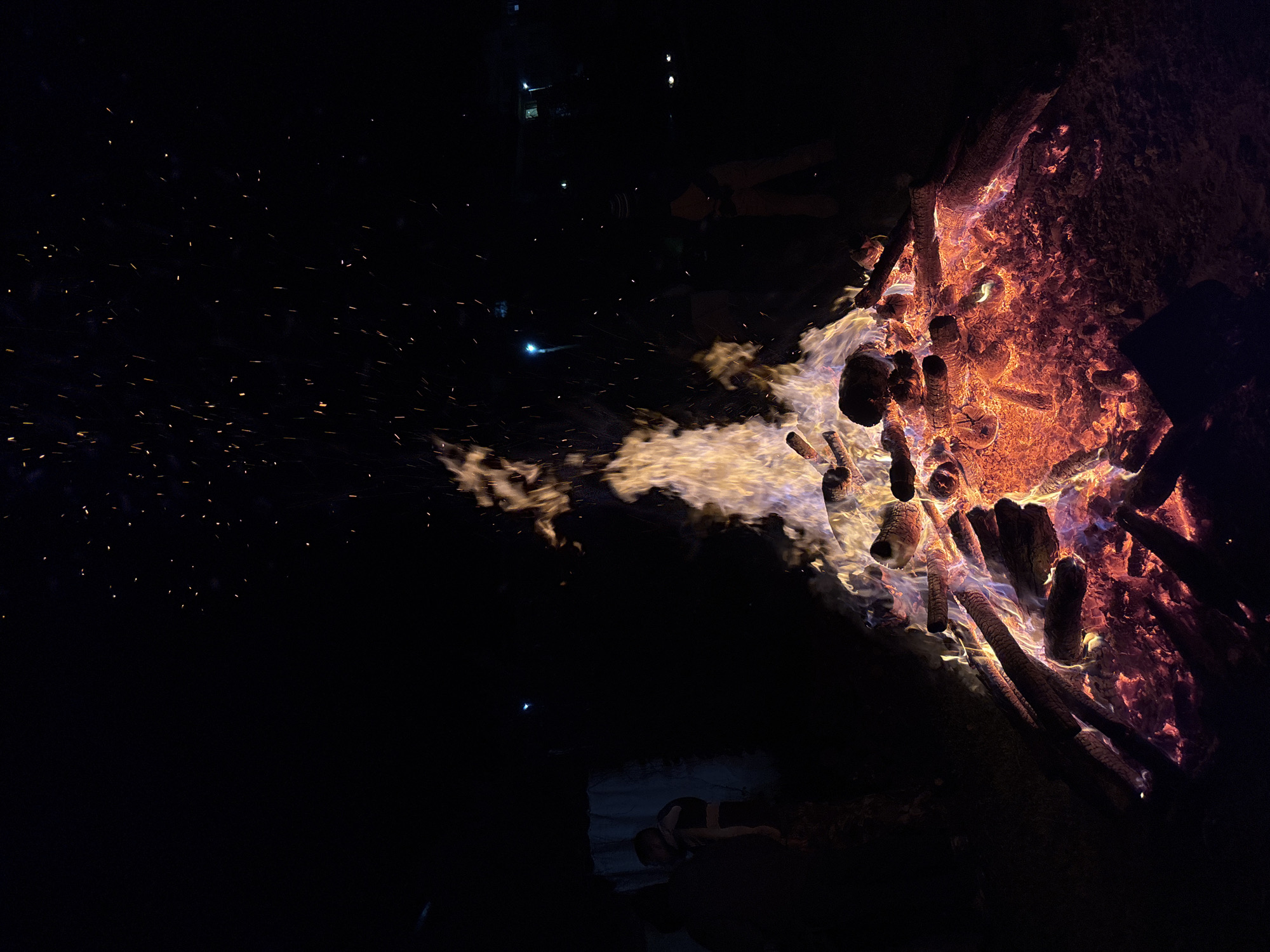 A nighttime view depicts a large bonfire constructed from numerous logs and branches burning intensely. The fire is positioned on the right side of the frame, with flames and glowing embers visible. Sparks from the fire rise into the dark surrounding space, particularly towards the left side of the image. The lower portion of a figure is barely visible in the bottom of the image on the lower right of the fire, seemingly lying down or crouching near the fire's edge. In the distance behind the fire, indistinct shapes suggest the presence of structures and individuals, possibly illuminated by faint light sources. The image has poor visibility in the distance. The location cannot be verified as Zlatograd, Bulgaria, from the visual data provided.