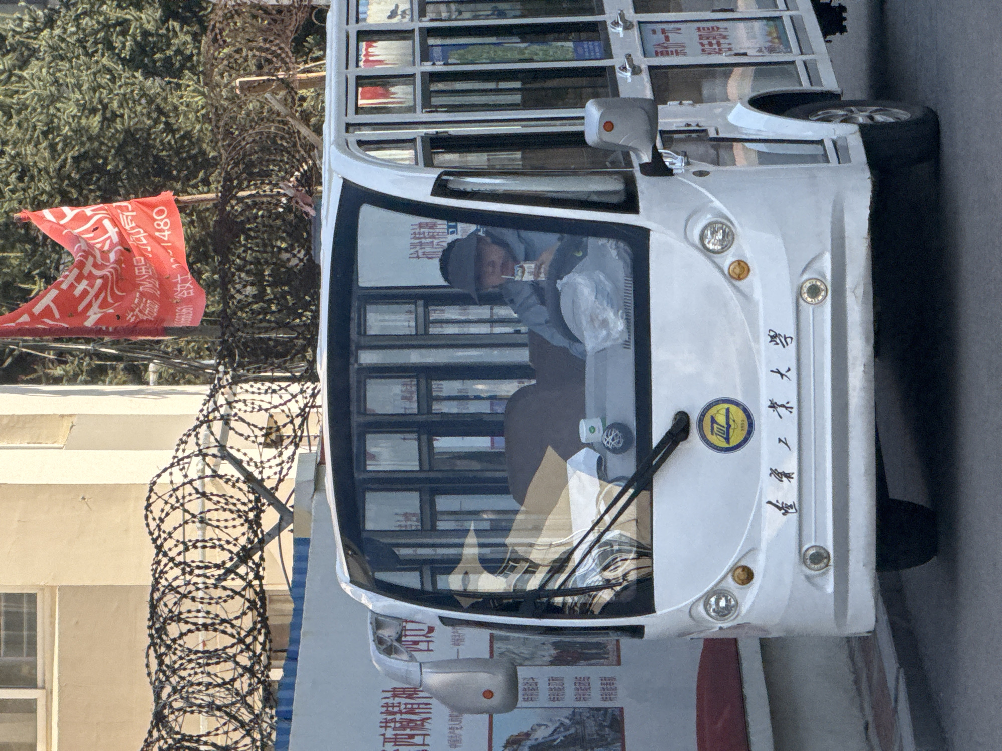 The image, captured in Zhongtun, China, depicts a white shuttle bus parked outdoors under bright daylight. A person is visible in the driver's seat, wearing a dark cap and a light-colored jacket, holding a carton with a straw to their mouth, appearing to drink. A white plastic bag is placed beside them.
The front of the bus displays headlights, a turn signal, and windshield wipers. Below the windshield, a circular yellow and blue logo with the year "1953" is visible, alongside Chinese characters written vertically that read "福建工业大学" (Fujian University of Technology).
To the left of the bus, a red flag with white Chinese text waves prominently. Visible phrases on the flag include "西藏精神" (Tibetan spirit), "中国共产党人的能" (The ability of Chinese Communists), and numbers such as "+ 12~1480". Some of this text, specifically "西藏精神" and "中国共产党人的能", also appears on the lower side panel of the bus, below the side mirror.
The background features dense green foliage behind the flag. A prominent, tightly coiled barbed wire fence (razor wire) stretches vertically, acting as a barrier. Beyond the fence, a light-colored building with windows is partially visible. The overall setting suggests a campus or institutional environment, possibly with restricted access given the presence of the barbed wire, and hints at political or organizational affiliations through the flag and bus text.