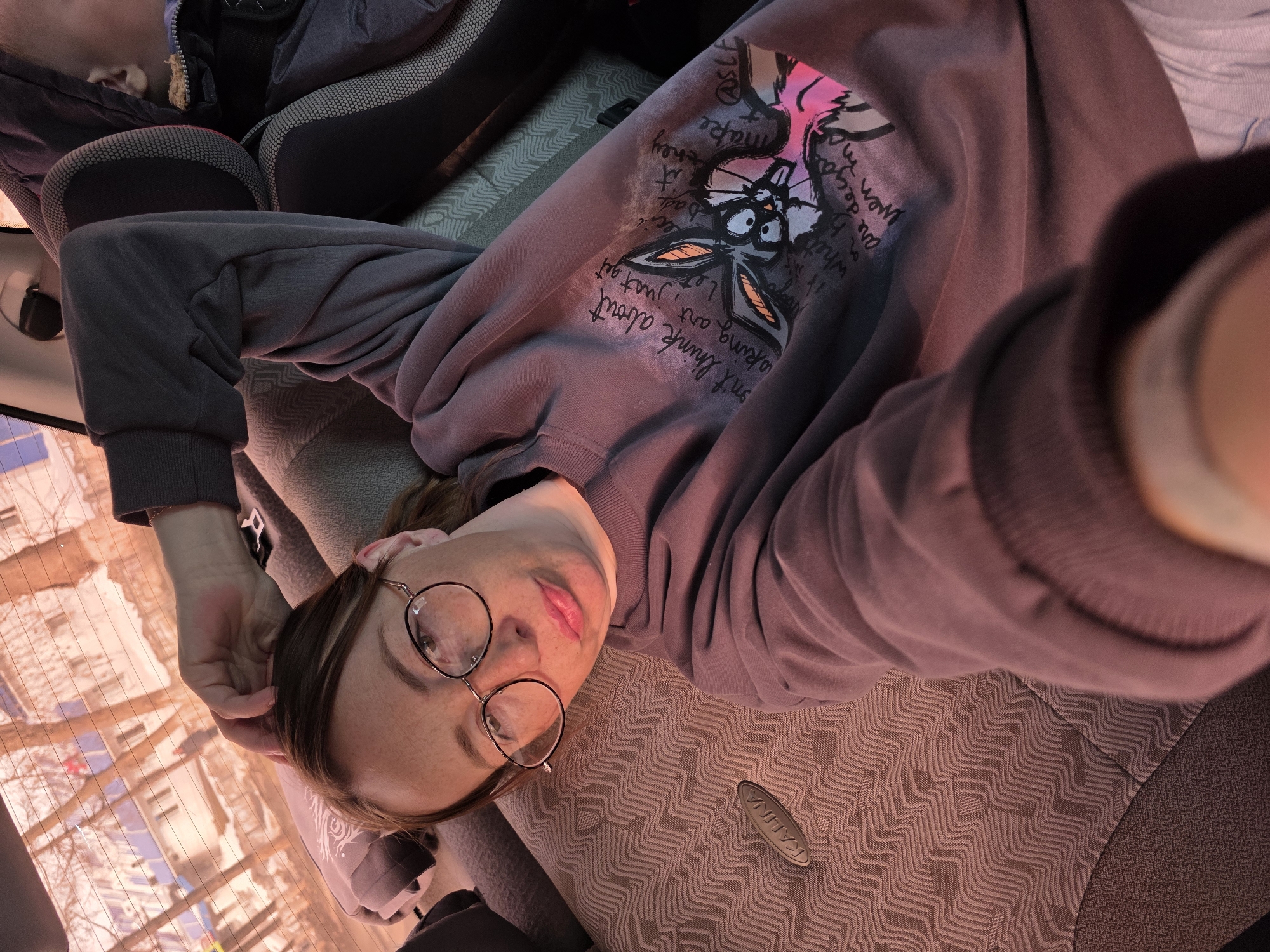A woman wearing round glasses and a gray sweatshirt lies on a car seat with a patterned fabric. She looks towards the camera with her head tilted. The sweatshirt has text and a cartoon design on it. Through the car window, buildings are visible.