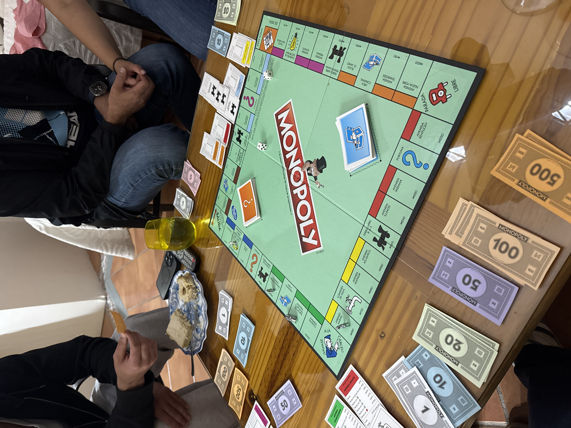 A group of people are playing Monopoly on a wooden table. The game board is centered on the table with dice on the "Nada Más" space, and Monopoly money is organized beside it in denominations of 1, 10, 20, 50, 100, and 500. Property cards are stacked next to the board. A glass and a plate of food are to the side. Two people are visible on the left side of the table, while another is visible on the right. The location cannot be determined from the image.